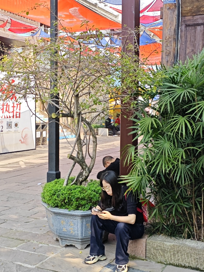A woman in jeans and a black shirt sits on a low stone wall, looking at her phone. She wears glasses and sneakers. Behind her, a man in black is leaning against a tree in a planter. Lush greenery surrounds them, with a taller bush to the right and smaller shrubs in the planter. Overhead, colorful canopies create a festive atmosphere. A poster is visible in the background.