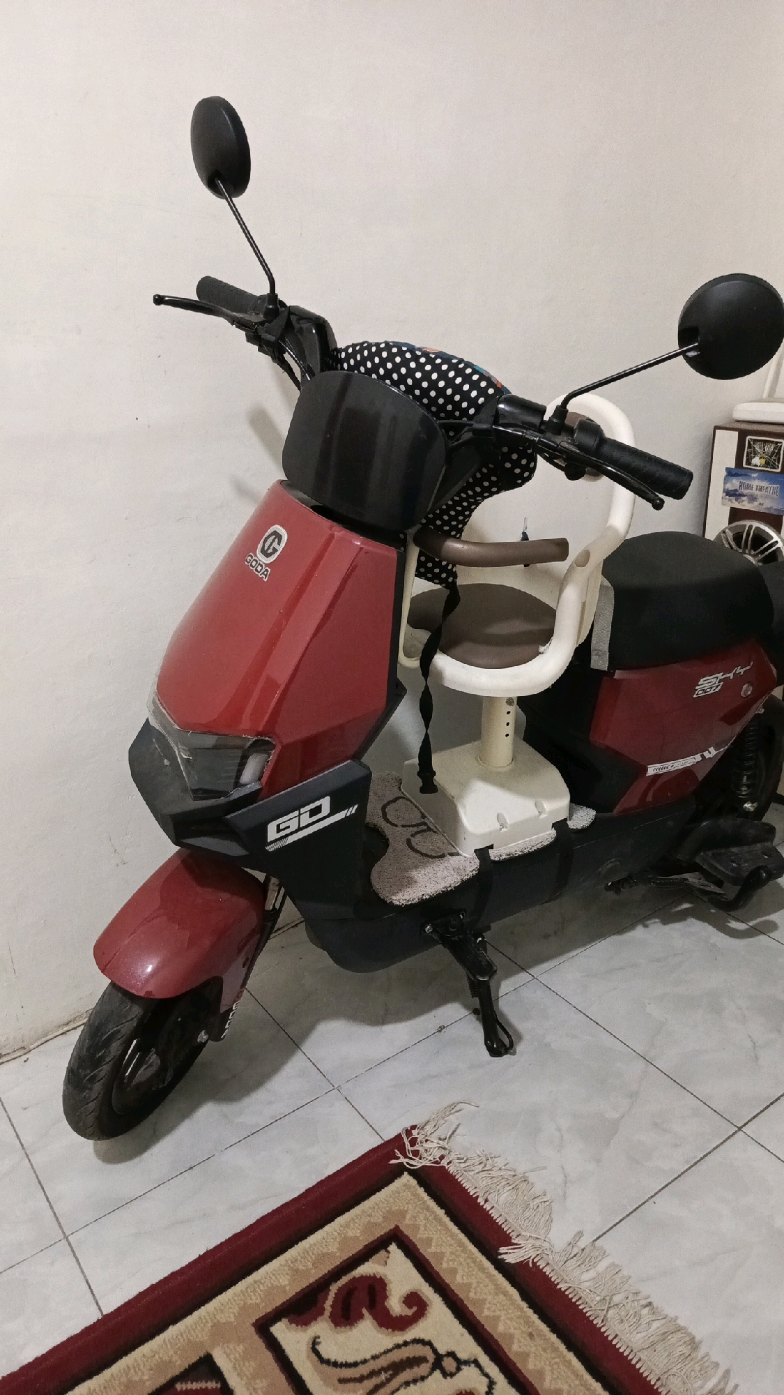 The image shows a red electric scooter with a child seat attached, parked indoors. The scooter has the "GODA" brand logo on the front. The environment appears to be a residential interior, possibly a garage or a room with tiled flooring and a white wall. A patterned rug is visible in the foreground. There are no people present in the image. The scooter is positioned at an angle, with its front wheel slightly turned. A small framed picture of a "HOME THEATRE" is visible on the wall in the background. The lighting suggests an indoor setting. The scooter is equipped with mirrors and handlebars. A patterned fabric with white polka dots is used as a cushion on the handlebars. The child seat is made of white plastic with a brown seat cushion and is secured with straps. The scooter has a kickstand deployed.