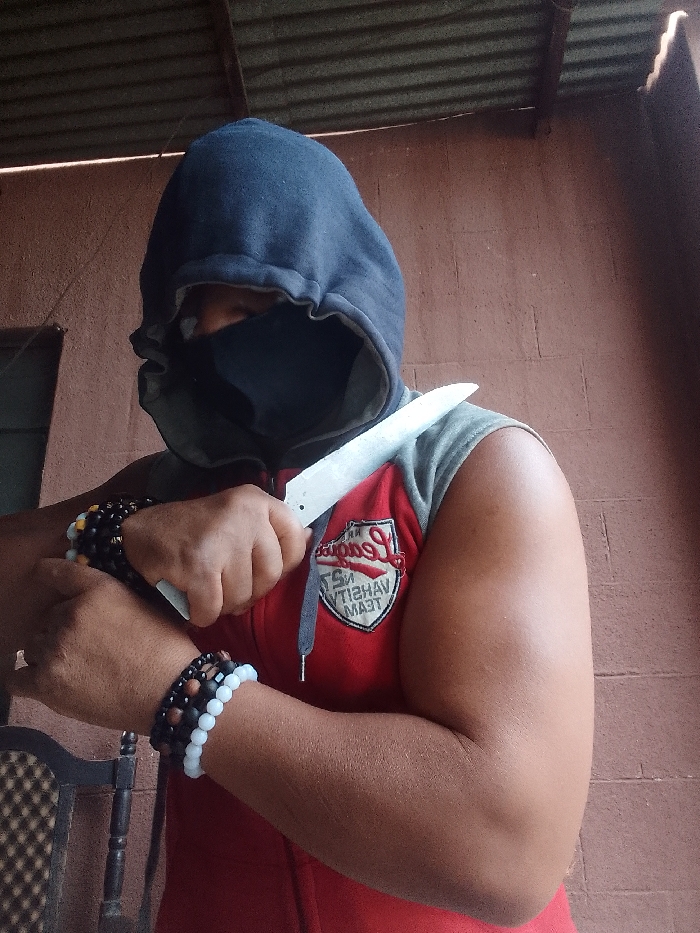 An individual is depicted indoors or in a semi-enclosed structure within Narayi, Nigeria. The person's identity is obscured by a dark blue hooded sweatshirt pulled over the head and a black face mask covering the nose and mouth. They wear a red sleeveless jacket with grey shoulder trim, featuring a white and grey embroidered emblem on the left chest with text partially visible as "COLLEGE...OF VARSITY TEAM." Both wrists display multiple beaded bracelets, including black, reddish-brown, yellow, and white/light blue beads. The individual holds a large, plain-bladed knife, possibly an unfinished blade, with both hands positioned horizontally across their upper torso. The background consists of a reddish-brown block wall, a corrugated metal roof supported by wooden beams, and a portion of a dark patterned chair on the left.