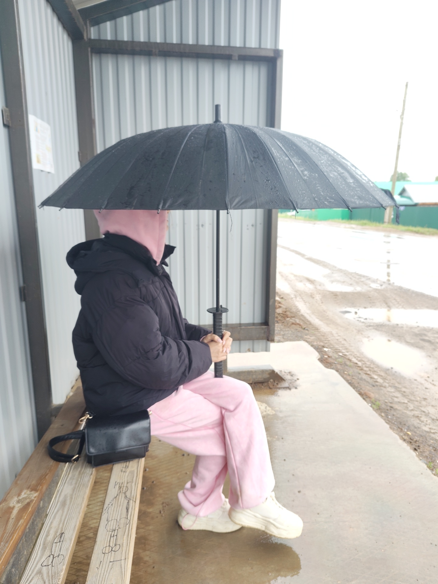 A person is sitting on a wooden bench under an umbrella in Foky, Russia. The person is wearing a black puffy jacket and light pink sweatpants and sneakers. A black purse sits on the bench next to them. The scene appears to be a bus stop or waiting area with a metal structure behind the bench. The ground is wet and there are puddles on the road visible in the background.