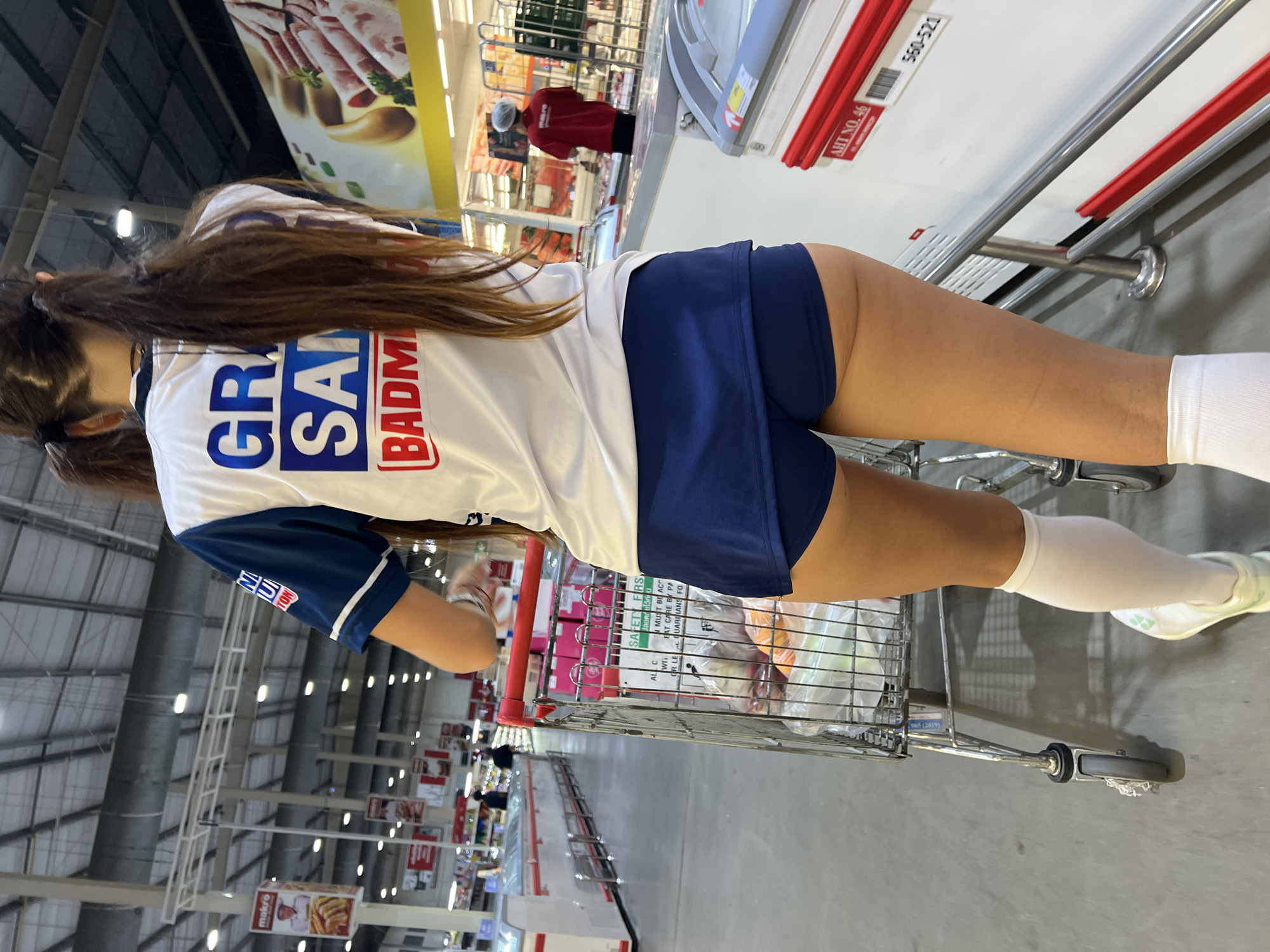 In this vertical, eye-level shot, a young woman with long brown hair is pushing a shopping cart in what appears to be a large warehouse-style store. She is wearing a white t-shirt with blue and red lettering that reads "GR SAN BADM" on the back and a dark blue skirt. She is also wearing white knee-high socks and athletic shoes. The shopping cart is filled with various items, some of which are in plastic bags.
In the background, the store is brightly lit with fluorescent lights and features high ceilings with exposed ductwork. Aisles of products are visible, and other shoppers can be seen in the distance. A large promotional banner with images of cured meats hangs from the ceiling. Text visible in the image includes "makro," "SAFETY FIRST," "MUST BE AC," "ALL C," "WI," "OR LE," "BAT CARE BY PA," "UARDIANS FO," "AHT NO. 46," and "560-521." The scene suggests a shopping trip in a commercial setting. The orientation of the photo is rotated 90 degrees counter-clockwise.