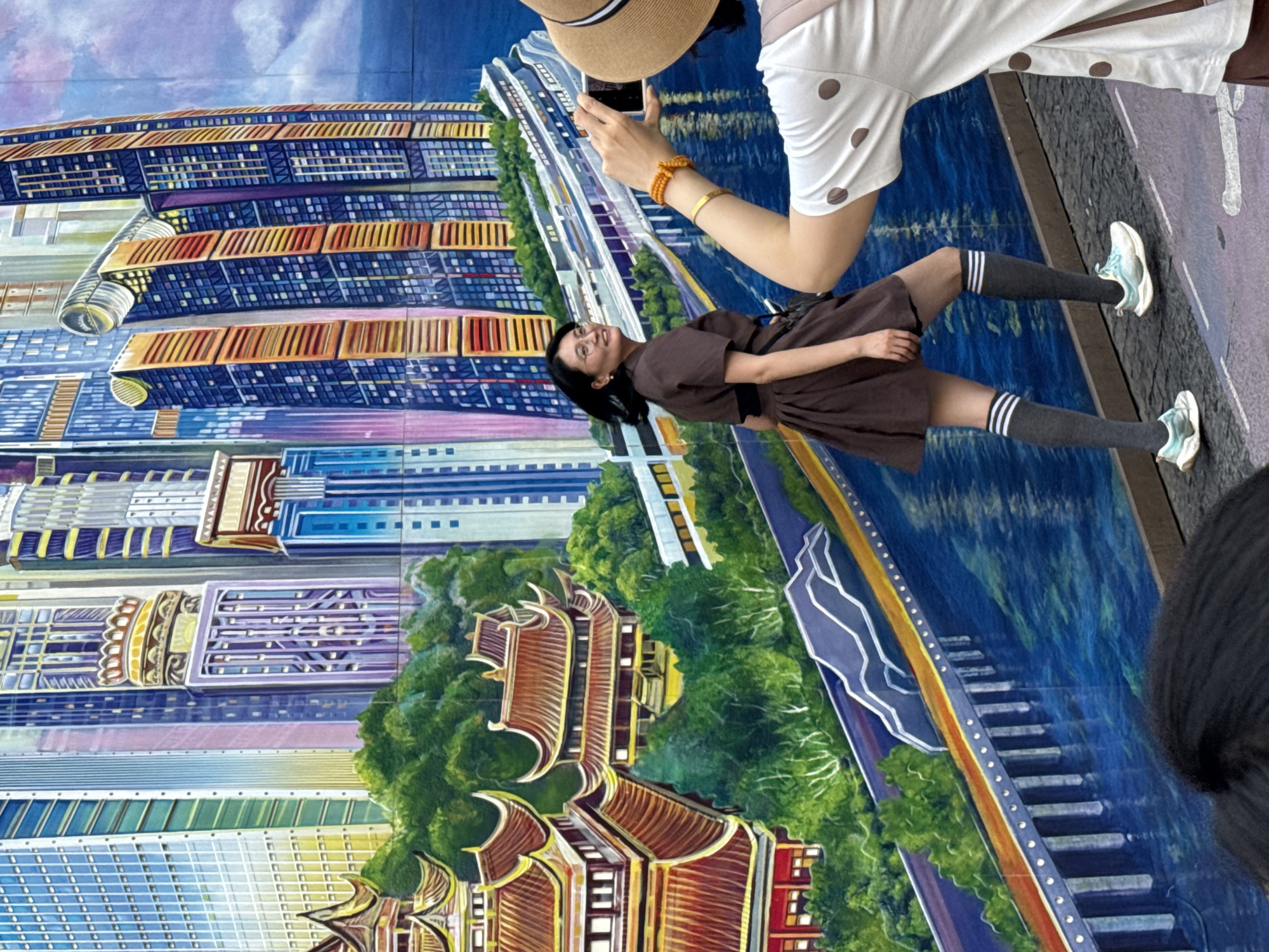 A woman in a brown dress and gray knee socks poses in front of a mural of Yuzhong District, China. A person wearing a hat and white shirt with brown spots stands nearby, taking a picture of the woman with a smartphone. The mural features various buildings and structures depicted in a stylized, colorful manner. A depiction of the Yangtze River and Jialing River flows horizontally across the backdrop.