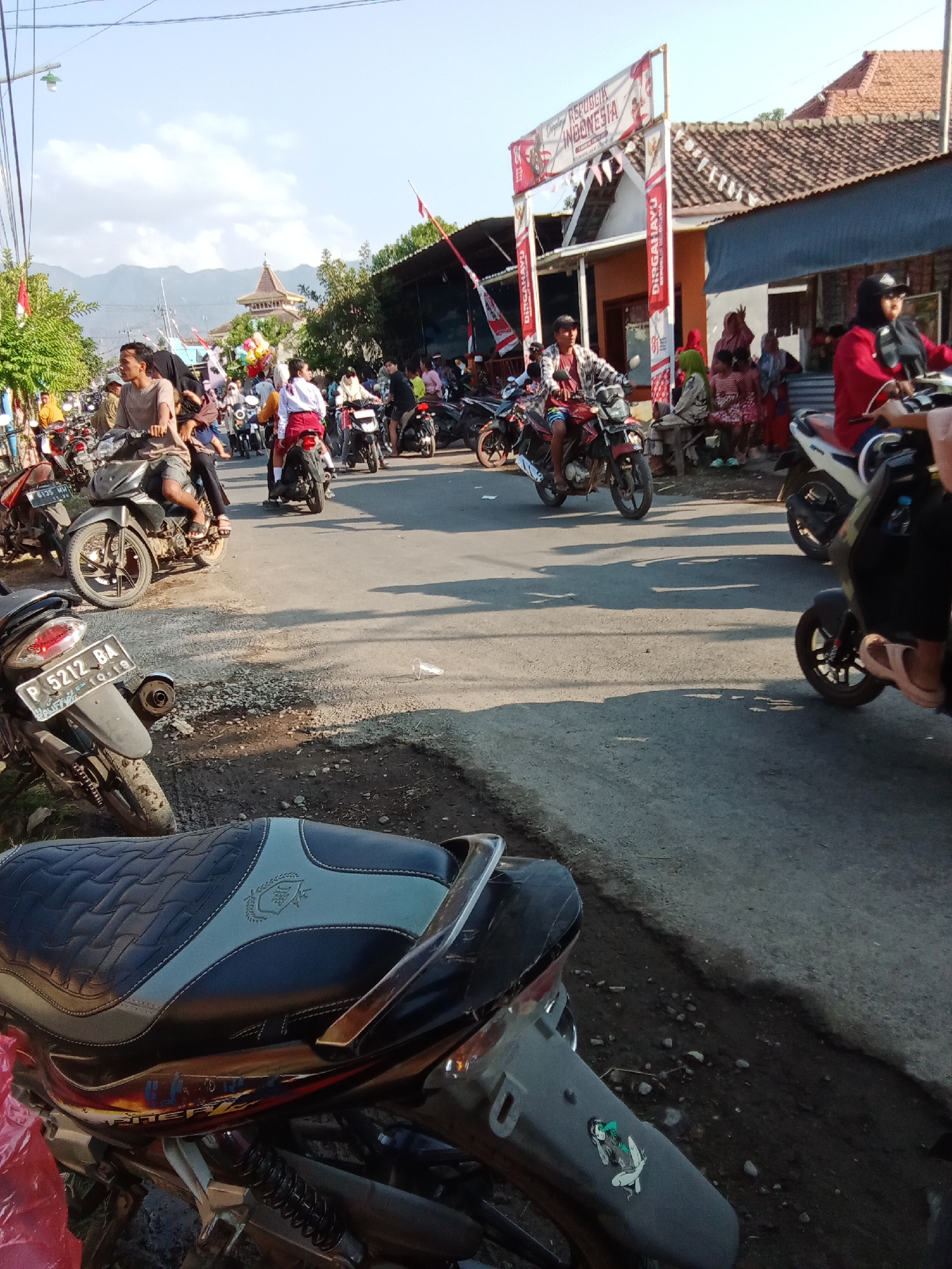 The image depicts a street scene in Bungatan, Indonesia, likely during a celebration, given the festive banners and the presence of many people. The street is paved and appears to be a main thoroughfare, with buildings lining one side. A group of people are riding and standing around motorcycles, suggesting a common mode of transportation and a community gathering. Some individuals are on the motorcycles, while others are gathered in small groups. The sky is clear and blue, indicating fair weather. In the background, a distinct architectural structure with a tiered roof is visible, potentially a religious building or a cultural landmark. There are also decorations and banners, including one with text that appears to be in Indonesian, celebrating "DIRGAHAYU REPUBLIK INDONESIA," which translates to "Long Live the Republic of Indonesia," indicating a national holiday or anniversary. The overall atmosphere appears lively and communal.