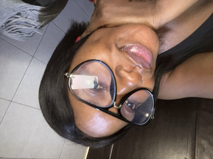 The image displays a close-up portrait of a woman, taken indoors in Dzhidu, Nigeria. She has dark skin, dark, straight hair, and is wearing large, black-framed glasses with clear, reflective lenses that feature gold-colored accents. Her lips are glossy, and she looks directly towards the camera with a slight smile. Her right hand, showing a thumb with red nail polish, is positioned under her chin/jawline. She is wearing a dark-colored garment visible on her left shoulder and chest. The background is divided into two sections: a dark brown, possibly wooden, surface in the upper left, and a light grey tiled wall in the lower right. A grey, textured fabric object is partially visible in the bottom right corner of the frame.