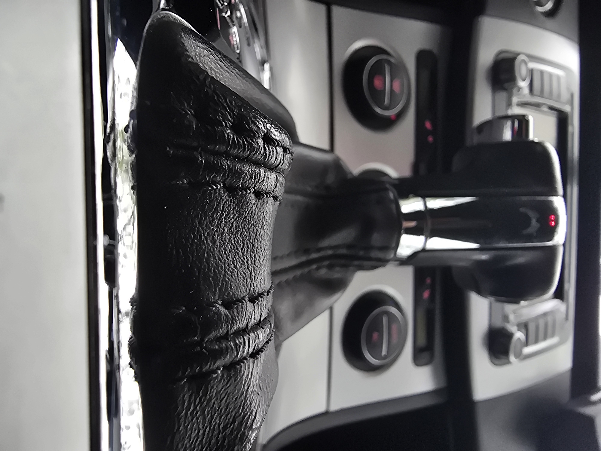 The image displays a close-up, low-angle view of the interior of a car, primarily focusing on the gear shifter and a section of the dashboard or center console. The main subject, a black gear shifter, is prominently featured in the foreground. It is made of a textured material, possibly leather or a synthetic equivalent, with visible horizontal stitching that adds to its tactile quality.
In the blurred background, a silver or grey control panel is visible, featuring two circular black knobs. Each knob displays a reddish-pink illuminated symbol, which appears to be a power or standby icon (a circle with a vertical line). Below the right knob, there are hints of additional controls or buttons, though these are indistinct due to the shallow depth of field. Shiny, chrome-like accents are present on the gear shifter, reflecting light from an unseen source. The overall aesthetic suggests a modern vehicle interior. No people are visible, and no specific activity is taking place. The image emphasizes the textures and design elements within the car's cabin, with the internal illumination on the controls suggesting the vehicle is powered on. No discernible text is visible.