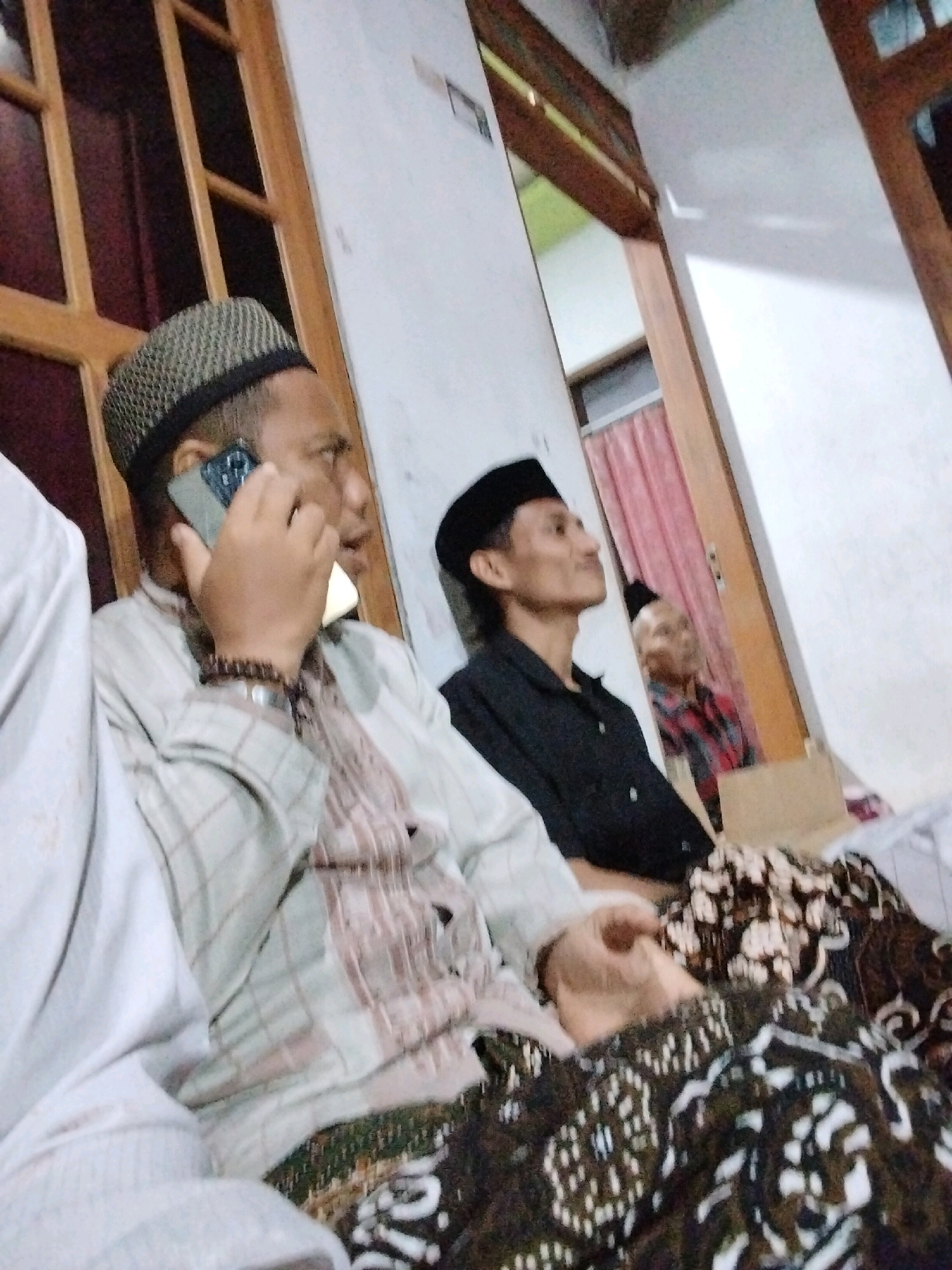The image depicts an indoor scene with several individuals, likely in a home setting in Tasikmadu, Indonesia. In the foreground, a man wearing a patterned skullcap and a checkered shirt is holding a smartphone to his ear and speaking, suggesting a phone conversation. He is seated, and a batik fabric is visible draped over his lap. To his right, another man wearing a black skullcap and a black shirt is looking away from the camera. Further in the background, a third man is partially visible, wearing a red and black checkered shirt and a skullcap. The environment appears to be a room with wooden doors and white walls. The lighting is dim, casting shadows and contributing to a somewhat blurred overall image quality.