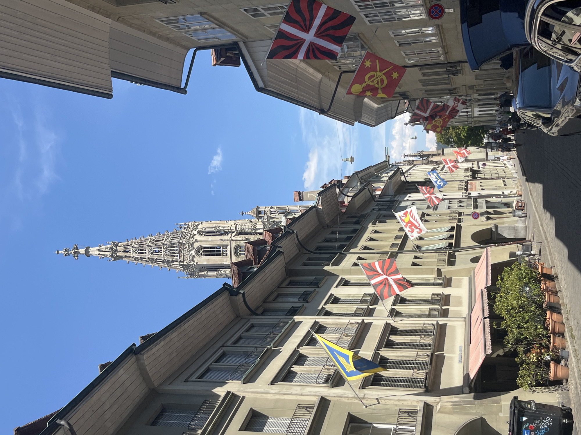 The image depicts a narrow street in Bern, Switzerland, with a prominent Gothic cathedral spire visible in the background. The street is lined with multi-story buildings with traditional architecture, featuring arched ground floors and windows with shutters. Several flags are displayed from the buildings, including the flag of Switzerland (a white cross on a red background) and various cantonal or civic flags with different designs. There are a few people walking down the street, and cars are parked along the side, with one with its trunk open. Potted plants are placed outside a building with awnings. The sky is clear and blue, indicating a sunny day. The overall scene suggests a historic European city with a vibrant atmosphere.