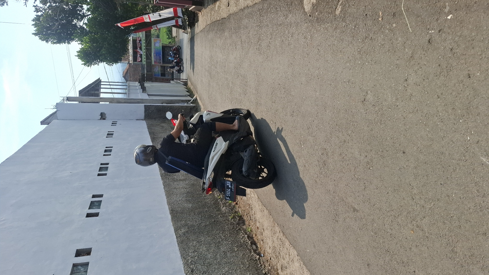 A person wearing a black shirt and helmet rides a white and black motorcycle on a paved road. The motorcycle has a license plate that reads "F 5924 XU". The person is positioned in the center of the frame, facing away from the camera and towards the right. They have their left arm on the handlebars and their right hand is out of frame. The motorcycle's shadow is cast on the road to the left.
To the left of the road is a textured gray wall with a series of rectangular openings, some of which appear to be glass. This wall is bordered by a lower, rougher gray surface.
To the right of the road, in the background, are several buildings with pitched roofs. A white structure with pillars and a covered roof is visible. Further down the road, there are more buildings and trees. Red and white flags are mounted on poles on the right side of the road. The sky is clear and blue, with some clouds. The overall scene suggests a sunny day in an outdoor urban or semi-urban environment in Cilaku, Indonesia.
