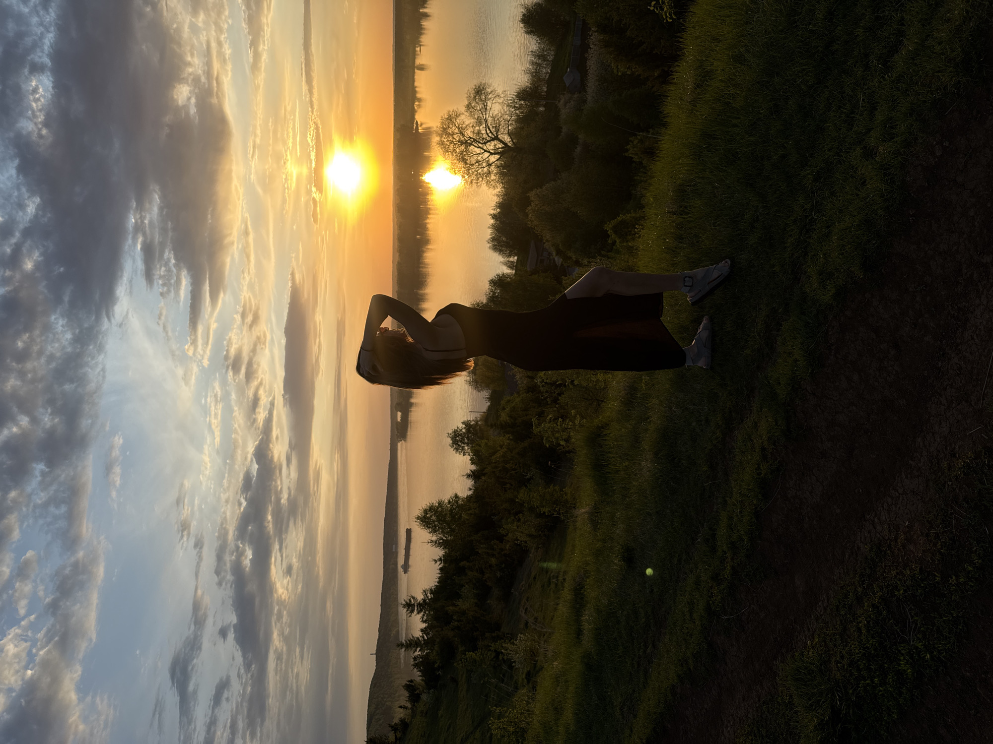A sideways view captures a woman lying on a grassy bank. She is facing the sky with her arms raised above her head. Her hair is spread out behind her. She wears a dark dress and light-colored sneakers.
Behind her is a body of water reflecting the setting sun. The horizon line is punctuated by trees and vegetation on the opposite shore. The sky is filled with clouds, some illuminated by the golden light of the sunset.
In the distance, a structure resembling a tower is visible. The location is Ольховка, Russia. A barge is visible on the body of water.