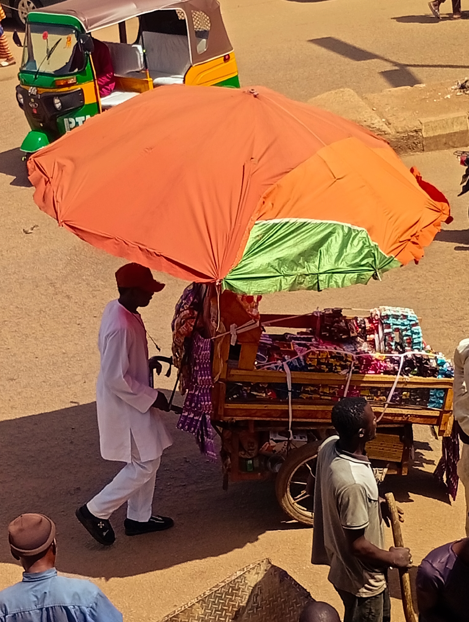 The image depicts a street scene in Bukuru, Nigeria, under bright sunlight. A vibrant orange umbrella, partially supported by a cart laden with colorful textiles, dominates the center of the frame. A man in a white tunic and red cap walks beside the cart, holding its handle. To his right, another man in a grey shirt stands holding a wooden stick. In the background, a green and yellow auto-rickshaw is parked, its passenger seat empty. The ground is a dusty, sandy surface, with a raised concrete curb visible. The overall impression is of a bustling market or roadside stall.
