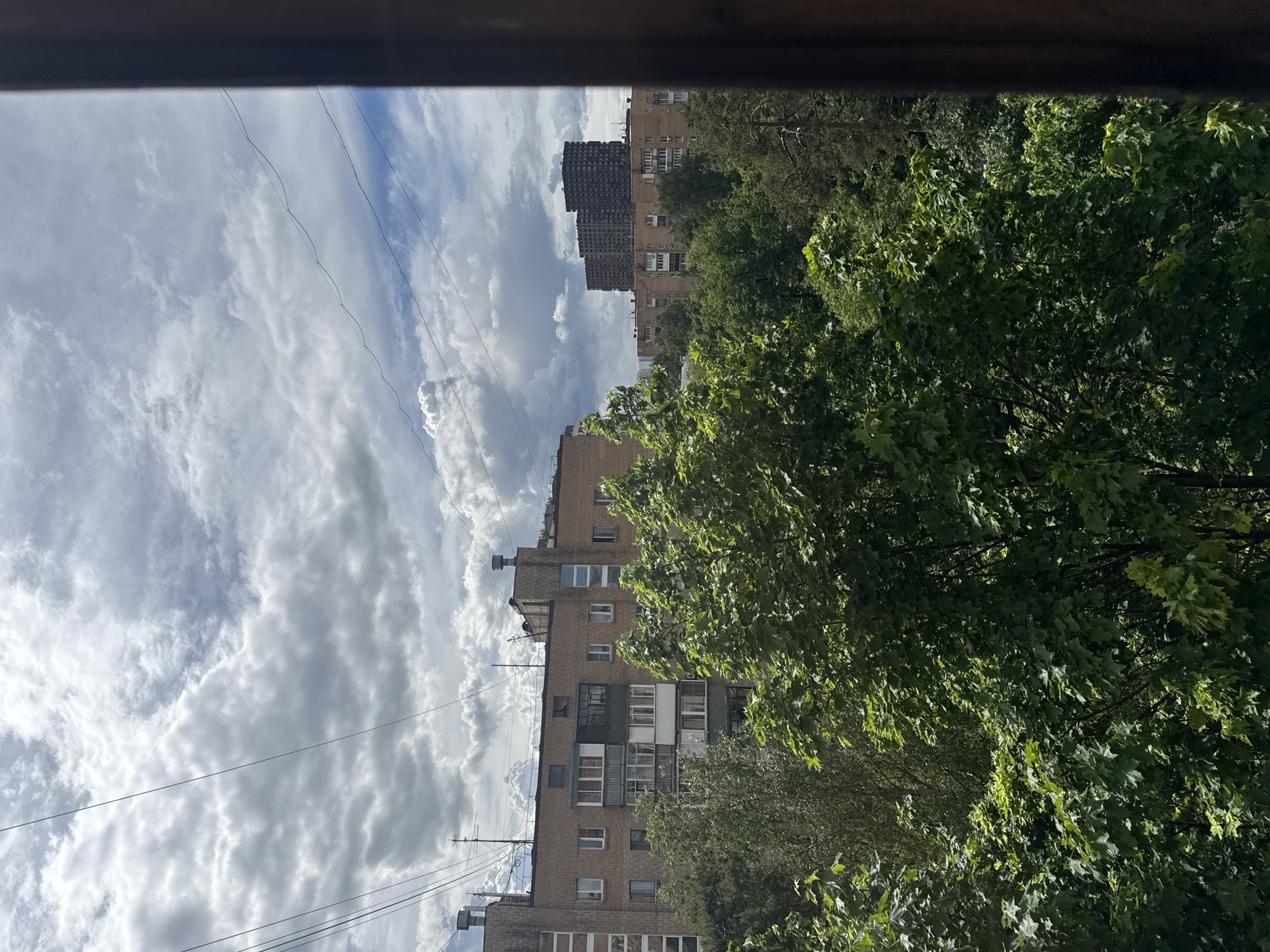 The image depicts a view from a window, possibly in Mytishchi, Russia, showing a cloudy sky, apartment buildings, and lush green trees.
The sky is filled with a mix of white and grey clouds, suggesting a potentially overcast day. Several apartment buildings of varying heights are visible, constructed of brick. The buildings appear typical of Soviet-era architecture.
A dense canopy of green trees occupies a significant portion of the right side of the frame. Power lines extend across the view.