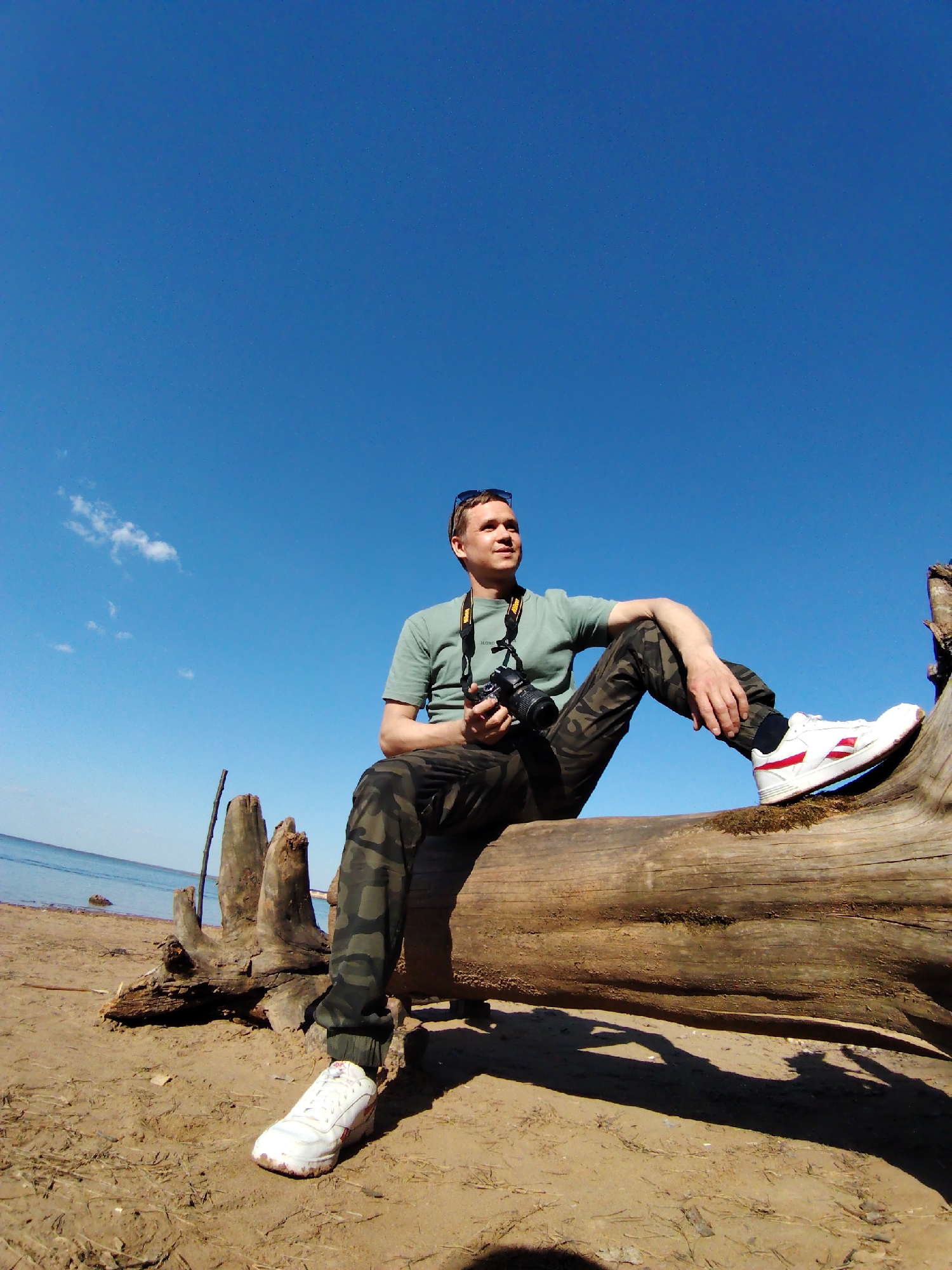A man is sitting on a fallen tree trunk on a beach under a bright blue sky with a few wisps of clouds. The beach is sandy, with remnants of driftwood and other debris. The man is wearing a green t-shirt, camouflage pants, and white sneakers. He is holding a camera and has sunglasses perched on his head. The scene appears to be set in Chaikovsky, Russia, as it's indicated in the query. The man is gazing towards the horizon. The light source is from above and to the right, casting a shadow of the log onto the sand. The ocean is visible in the background.