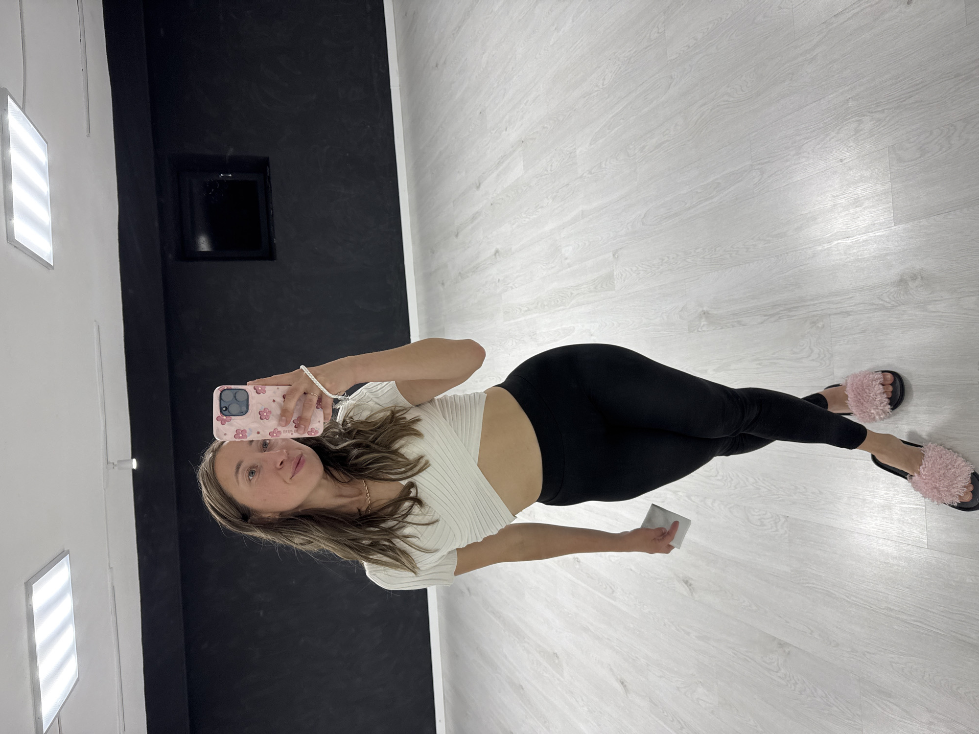 A young woman is captured in a selfie taken indoors, appearing to stand upright though the image is rotated 90 degrees clockwise. She has long brown hair and is wearing a light-colored, short-sleeved, ribbed crop top, black leggings, and pink fluffy slippers. A gold-colored necklace and a white beaded bracelet are also visible. She holds a pink smartphone with a floral pattern case in her right hand, which she uses to take the selfie, and a small, rectangular silver/grey object in her left hand.
The setting is a well-lit indoor space, possibly a studio or modern room. The floor features light-colored wooden planks with a distinct grain pattern. The walls are contrasting: a bright white wall on the left, where two illuminated rectangular light fixtures with horizontal strips are mounted, and a dark black wall on the right. A clear vertical line separates the two wall colors. The black wall contains a dark, recessed rectangular opening. The woman looks directly at the camera with a neutral to slightly smiling expression. Faint text, "little.", is visible on one of the flowers on her phone case.