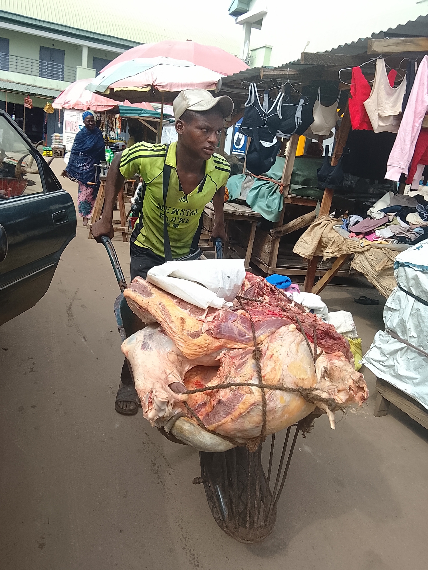 A young man wearing a lime green t-shirt and a light-colored baseball cap pushes a wheelbarrow laden with a large quantity of raw meat. The meat is tied with rope and appears to be from a whole animal. He is in a busy outdoor market in Akwanga, Nigeria, as suggested by the presence of stalls, umbrellas, and other people in the background. To his left, a dark-colored car is partially visible. In the background, a woman in a blue headscarf and a patterned pink and blue dress walks away from the camera. Various items, including clothing and other market goods, are displayed on shelves and tables in the stalls. The ground is paved.
