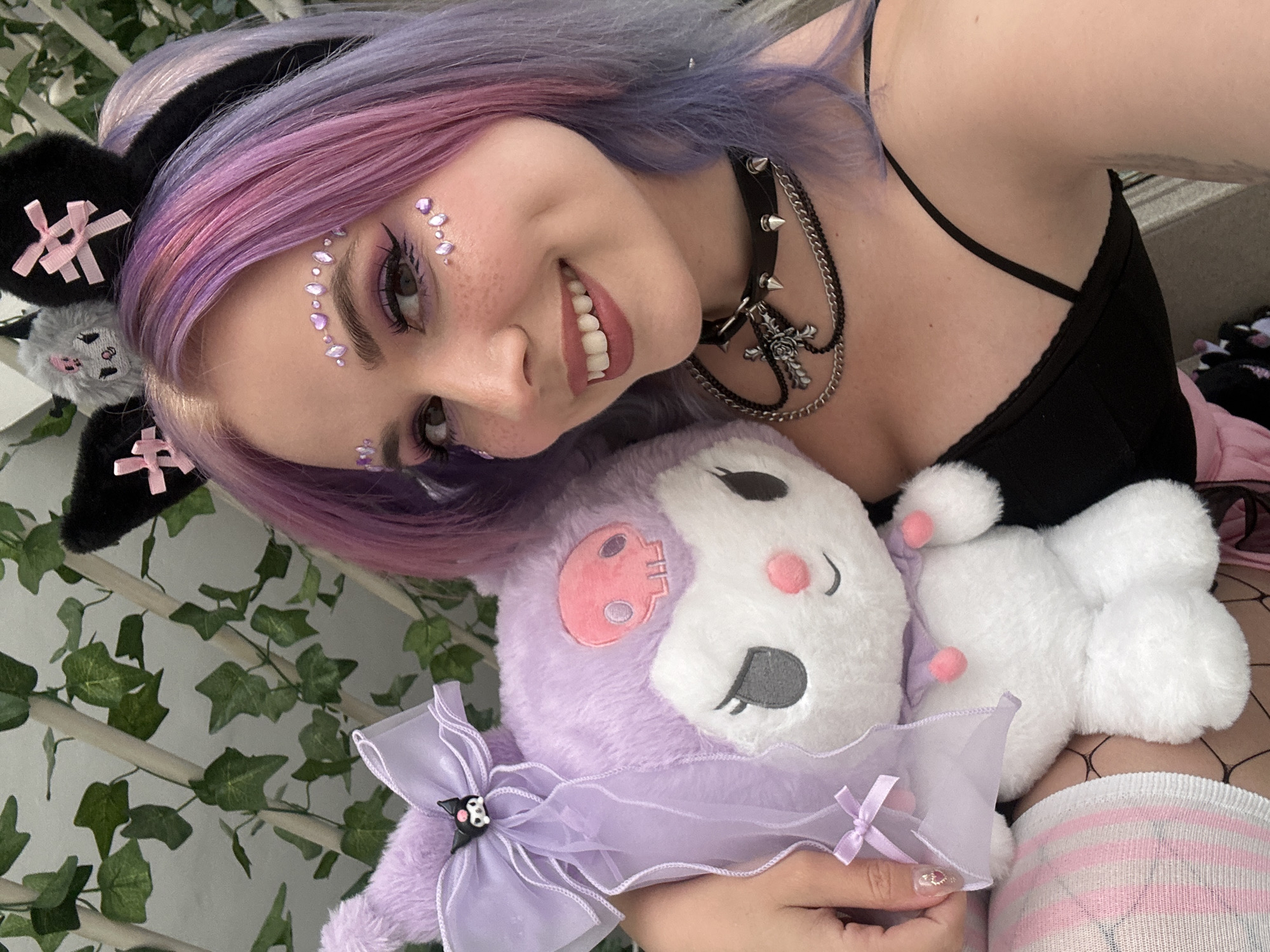 A person with purple-dyed hair and wearing a black tank top and a choker is holding a purple stuffed animal with a white face. The person has face gems and is smiling. They are wearing knee-high pink and white striped socks and fishnet stockings. The background includes green vines. The location cannot be determined based on the image provided.