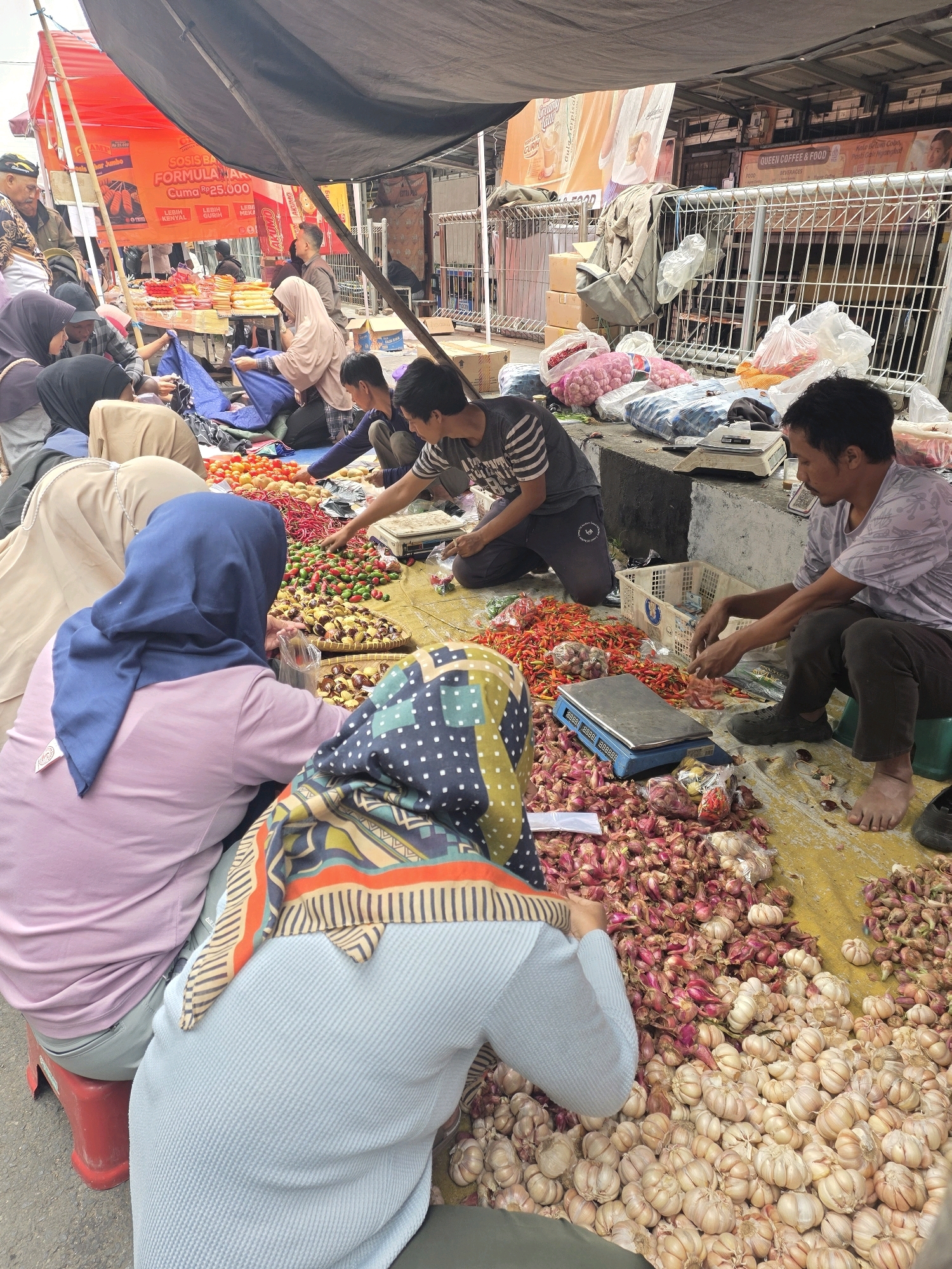 A group of people are gathered around a market stall in Bojongsoang, Indonesia, with a focus on agricultural produce. The scene takes place outdoors under a large black tarp, with a red tent visible in the background. The foreground is dominated by piles of shallots and garlic, with a blue digital scale placed on the yellow ground cover. Several individuals, appearing to be vendors and customers, are kneeling or sitting around the produce, some engaged in handling or weighing the items. One person in the foreground wears a light blue headscarf and a blue and white patterned scarf, and another wears a pink shirt and a blue headscarf. In the mid-ground, a man in a striped shirt is sorting through small red chilies. Behind them, other market activities are suggested, with more people and stalls partially visible. The overall impression is of a busy, local food market.