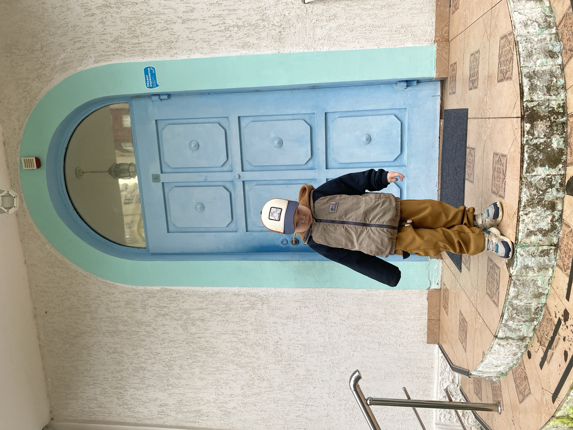 A young boy stands in front of a blue door framed by a light-blue arch. The door is comprised of six square panels, each adorned with a raised circular design at its center. The boy is wearing a brown jacket over a dark long-sleeved shirt, brown pants, and sneakers. He has a hat with a white panel on the front. The building's facade is white with a textured surface. The entrance is decorated with patterned tiles, and a metal railing is visible on the steps. A small sign with Russian text is attached to the door frame. This scene appears to be from Sochi, Russia.