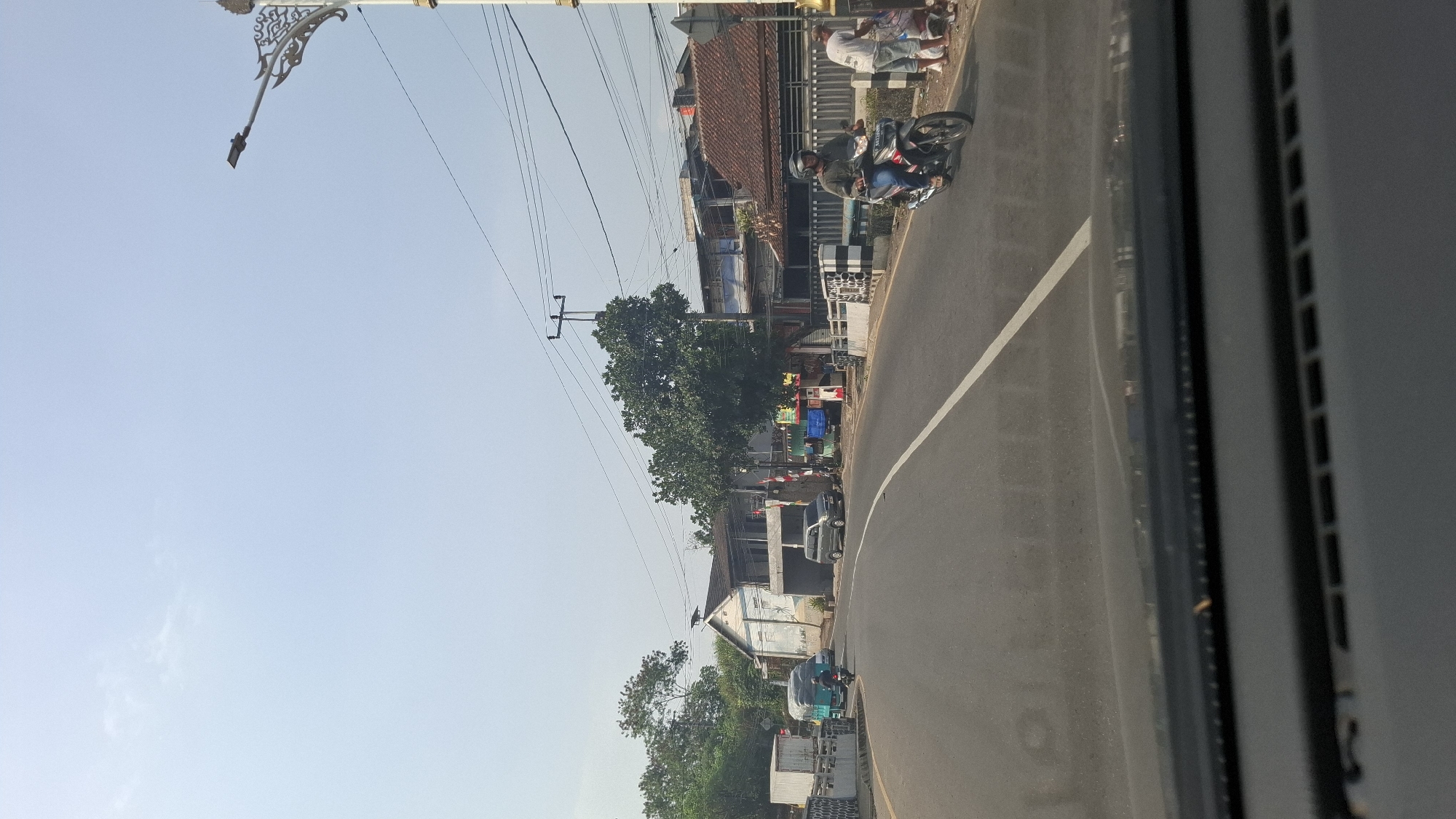 The image captures a street scene in Cilaku, Indonesia, likely during daylight hours with a clear sky. The perspective is from inside a vehicle, looking forward through the windshield. A paved road dominates the foreground, with a white line marking the lane.
On the right side of the road, a man is riding a red and black motorcycle, wearing a helmet. Next to him, on the sidewalk, two individuals are standing. One appears to be a woman wearing a headscarf and a patterned dress, while the other is a person in a white t-shirt and shorts.
Along the left side of the road, there are buildings with varying rooflines, some appearing to be shops or residences. A light blue truck with a green tarp covering its cargo is visible further down the road. In the distance, trees and vegetation line the sides of the street. A decorative street lamp with intricate metalwork stands on the right side of the image. The overall environment suggests a developing urban or semi-urban area.