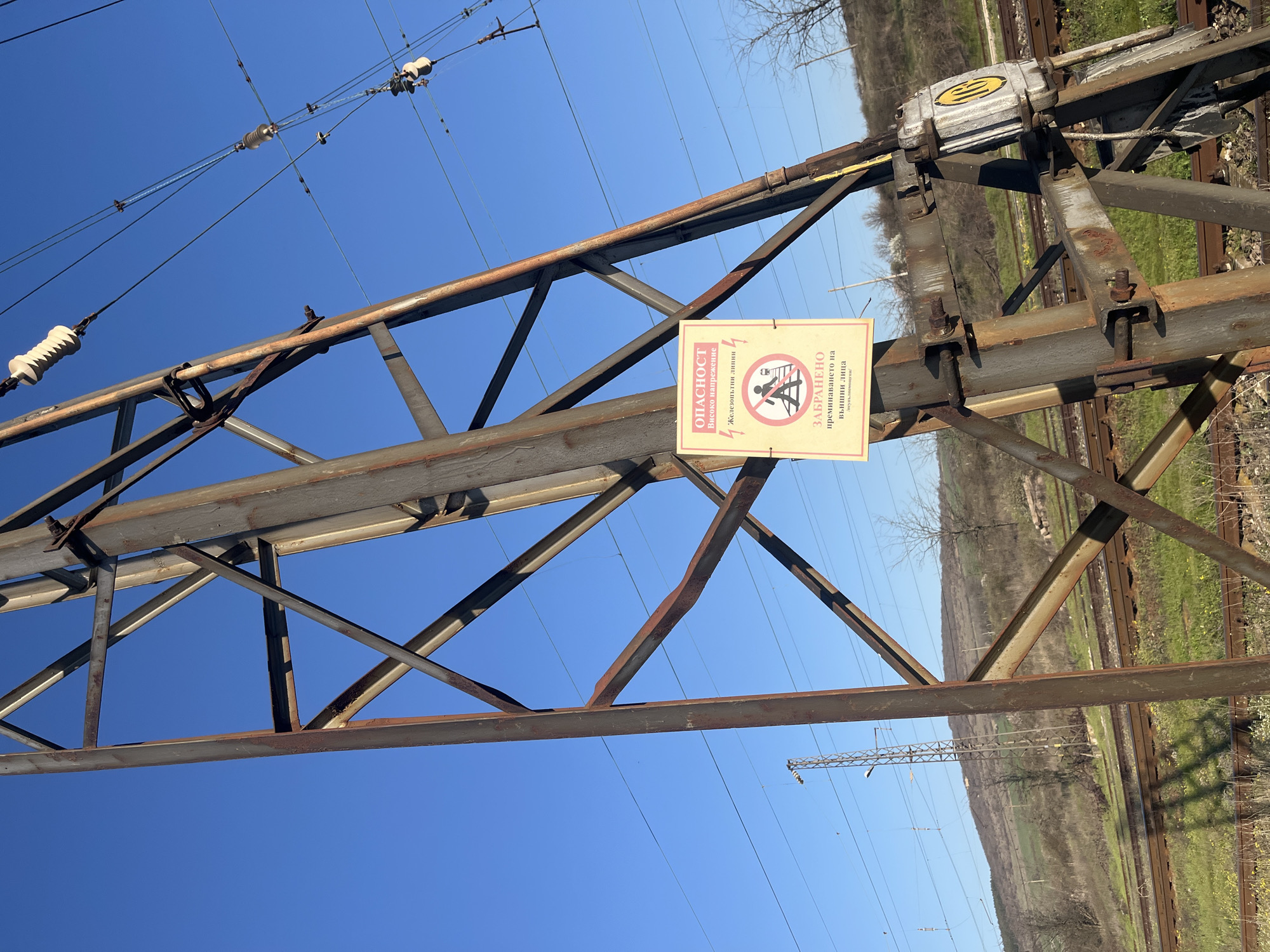 The image depicts an overhead power line structure in Lozarevo, Bulgaria. The structure is made of a rusty, metal frame with triangular reinforcements. A warning sign in Bulgarian is attached to the frame, cautioning about high voltage and prohibiting unauthorized access. High-voltage power lines are strung above the structure. In the background, railroad tracks run parallel to the power line, and there is vegetation on the opposite side of the tracks. The sky is clear and blue.