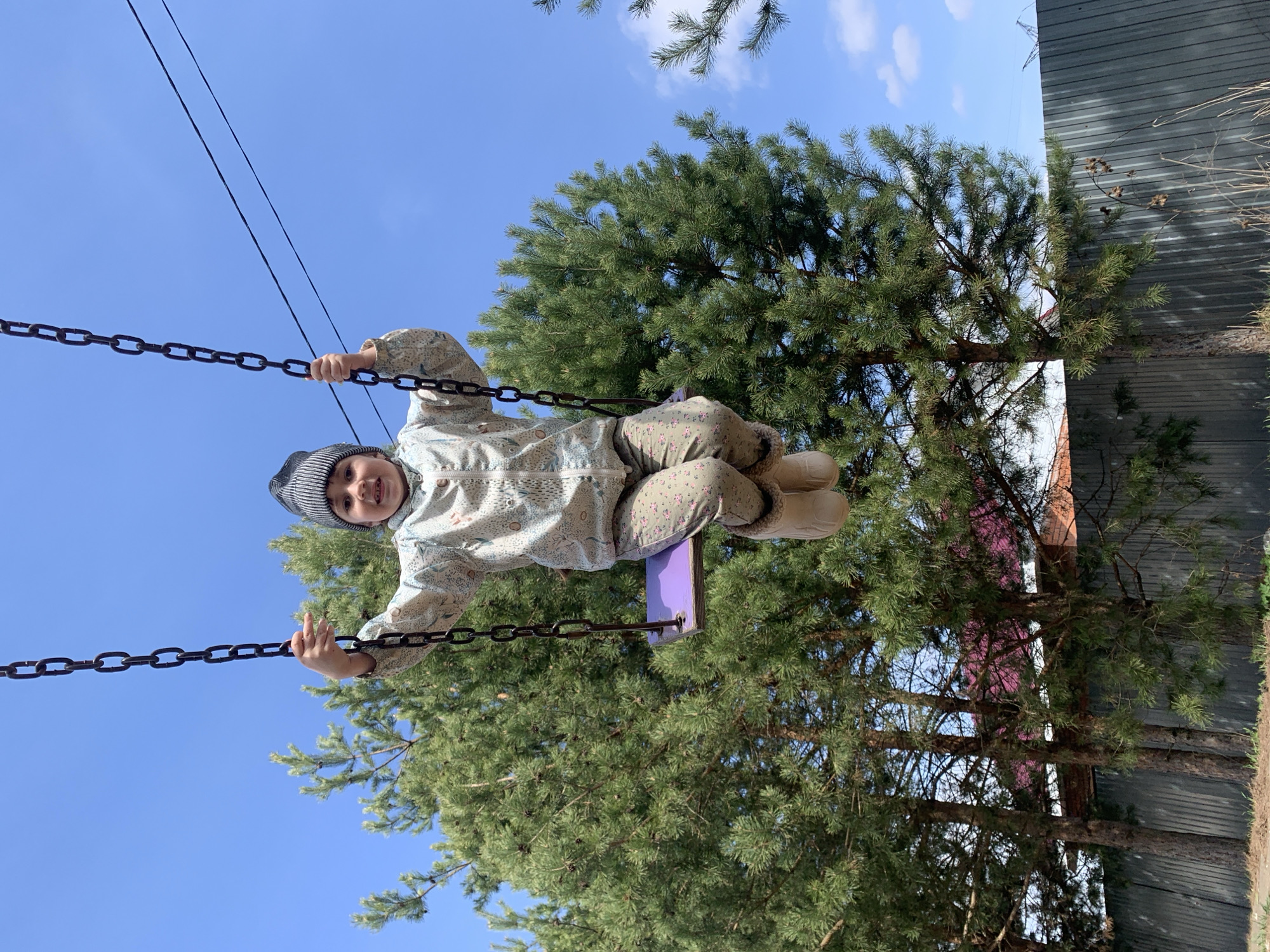 A child is on a swing in an outdoor setting in Chaikovsky, Russia. The child wears a patterned jacket and pants, boots, and a striped hat. They hold the swing chains. A pine tree is behind the swing. A section of corrugated metal fence and partial view of a building are in the background. The sky is blue with few clouds.