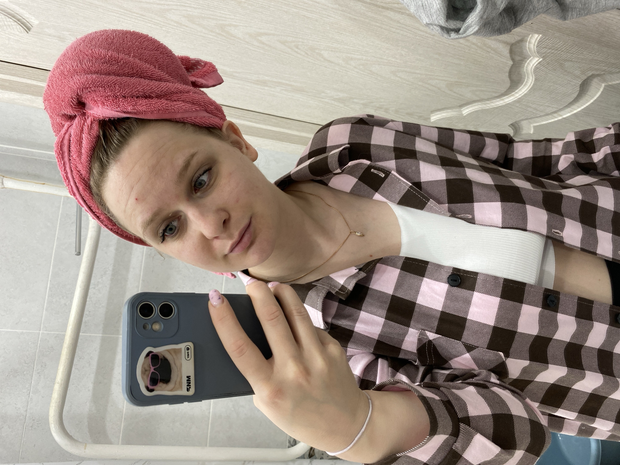 A woman is taking a selfie in what appears to be a bathroom. She has a pink towel wrapped around her head and is wearing a pink and brown plaid shirt over a white tank top. She is holding a blue smartphone with a pug sticker on the back. The background includes a white tiled wall, a white towel rack, and a glimpse of a light-colored surface with decorative molding.