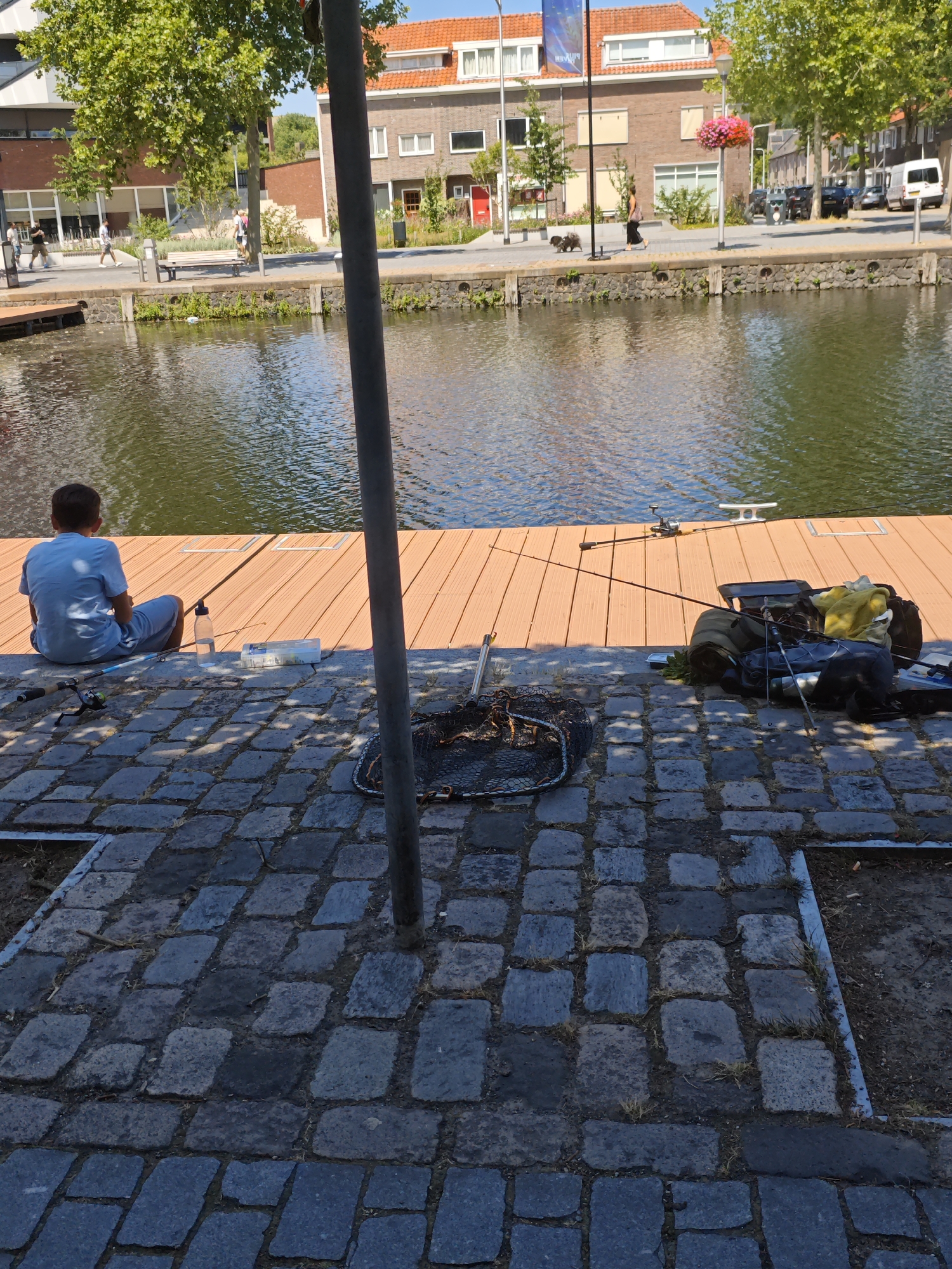A person is fishing by a canal in Tilburg, Netherlands. The scene depicts a person sitting on a wooden dock facing the water, with fishing equipment nearby. The canal is calm and reflects the surrounding buildings and trees. The area is paved with cobblestones in the foreground, and the background shows residential buildings and trees. The weather appears to be sunny and warm.