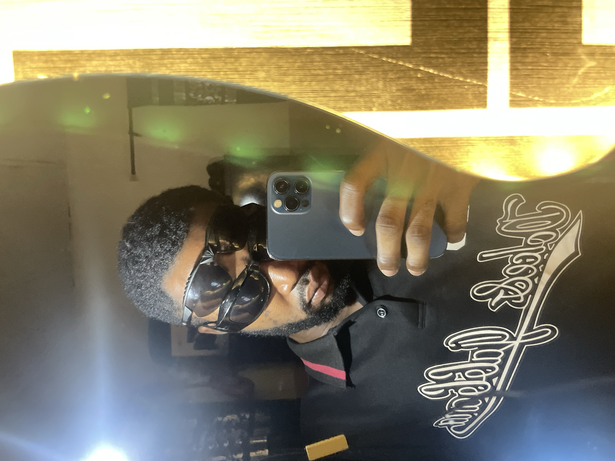 This image captures a Black male taking a mirror selfie in an indoor setting in Lokoja, Nigeria. The subject wears a black polo shirt featuring a red striped collar and white script lettering on the chest that reads "Lokoja" and a second word, appearing to be "Dodge." He has short, dark, curly hair and a trimmed beard. His eyes are obscured by two stacked pairs of dark sunglasses. The subject holds a dark gray/blue smartphone, identifiable by its triple-lens camera array, in his right hand to capture the reflection. A bright, circular light source is visible in the upper left portion of the mirror's reflection, causing significant glare. Several faint green, circular light artifacts are present on the mirror's surface, particularly on the right side. To the right of the subject, the reflection shows a textured, golden-yellow surface with vertical striations, possibly a wall hanging or furniture. A small, rectangular golden object is visible on the left side of the subject's chest area in the reflection.
