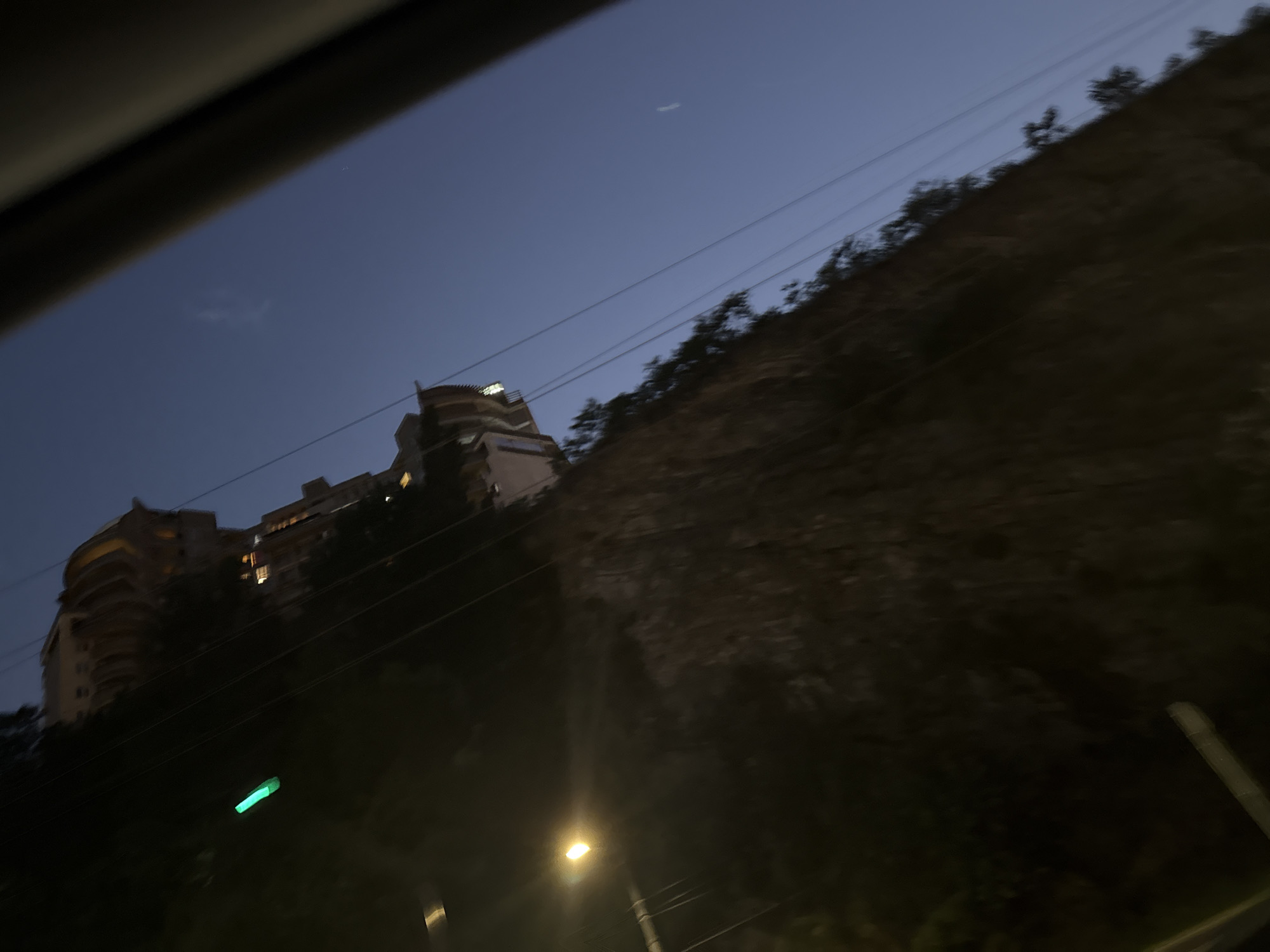 The image shows a view from a moving vehicle in Tbilisi, Georgia, during twilight. A steep, rocky incline is visible on the left, covered with sparse vegetation. Several power lines run diagonally across the frame. At the top of the incline, a multi-story building with illuminated windows is perched, appearing to be a residential or hotel structure. A bright, yellowish street lamp is visible on the left side of the image, casting a glow. A single green light is also present on the rock face, possibly a marker or indicator. The sky is a deep, clear blue, transitioning to darker tones as nightfall approaches. The perspective suggests the vehicle is traveling along a road that runs parallel to the incline.