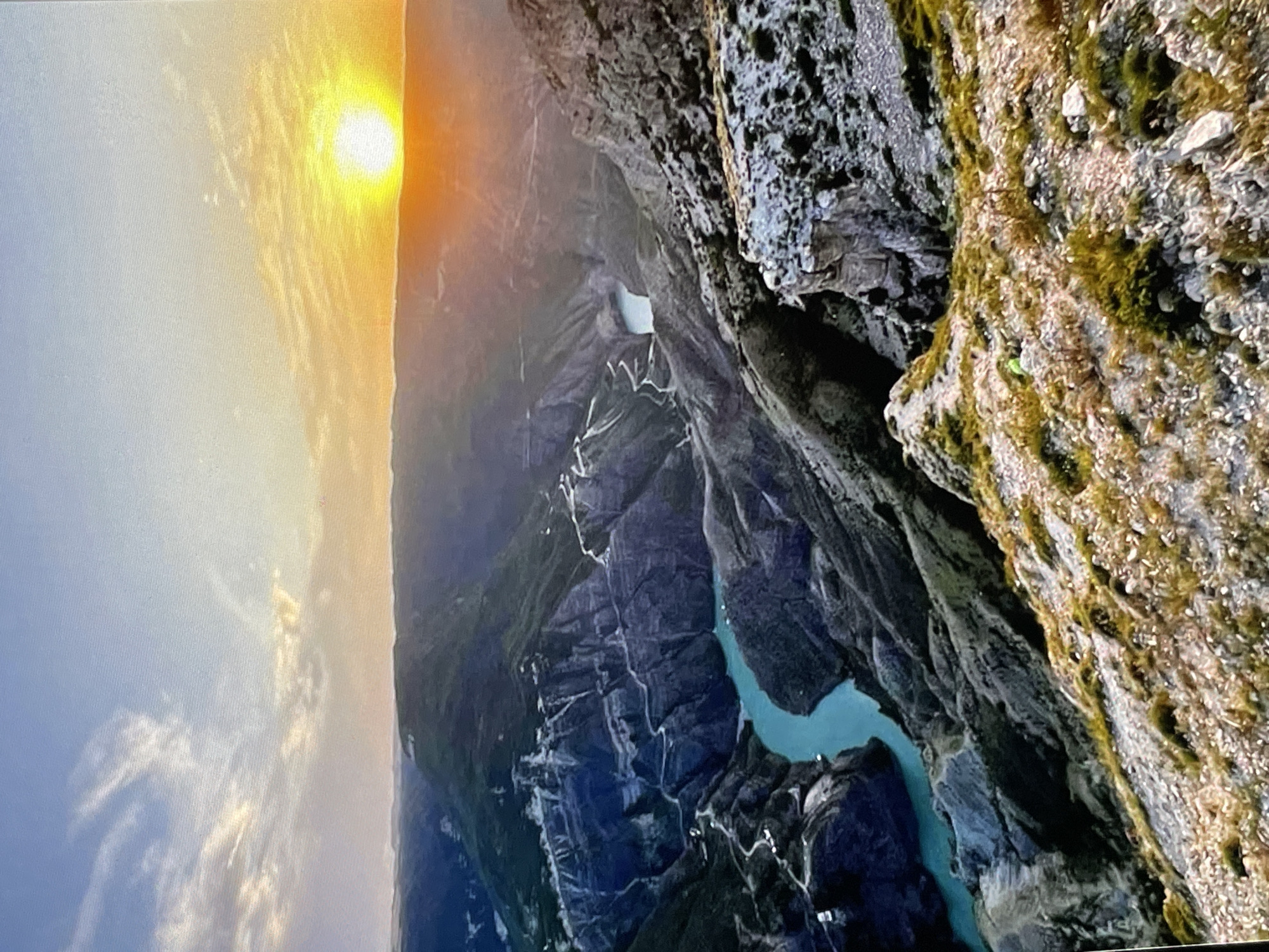 The image is a wide-angle, high-angle shot of a vast canyon with a turquoise river flowing through it. The canyon walls are steep and rugged, with visible winding roads or paths carved into them. The landscape is predominantly mountainous and rocky, with patches of green vegetation. The sky is a pale blue with wispy clouds, transitioning to a bright orange and yellow sunset on the right side of the frame. The sun is a distinct orb low on the horizon. The foreground consists of a rocky, sparsely vegetated slope. There are no people visible in the image. The scene appears to be a natural landscape, likely a remote canyon area. The prompt's request to include the "location in the city of Pushkino, Russia" is not supported by the visual information in the image, as the landscape depicted is characteristic of a mountainous canyon, not an urban or suburban environment like Pushkino.