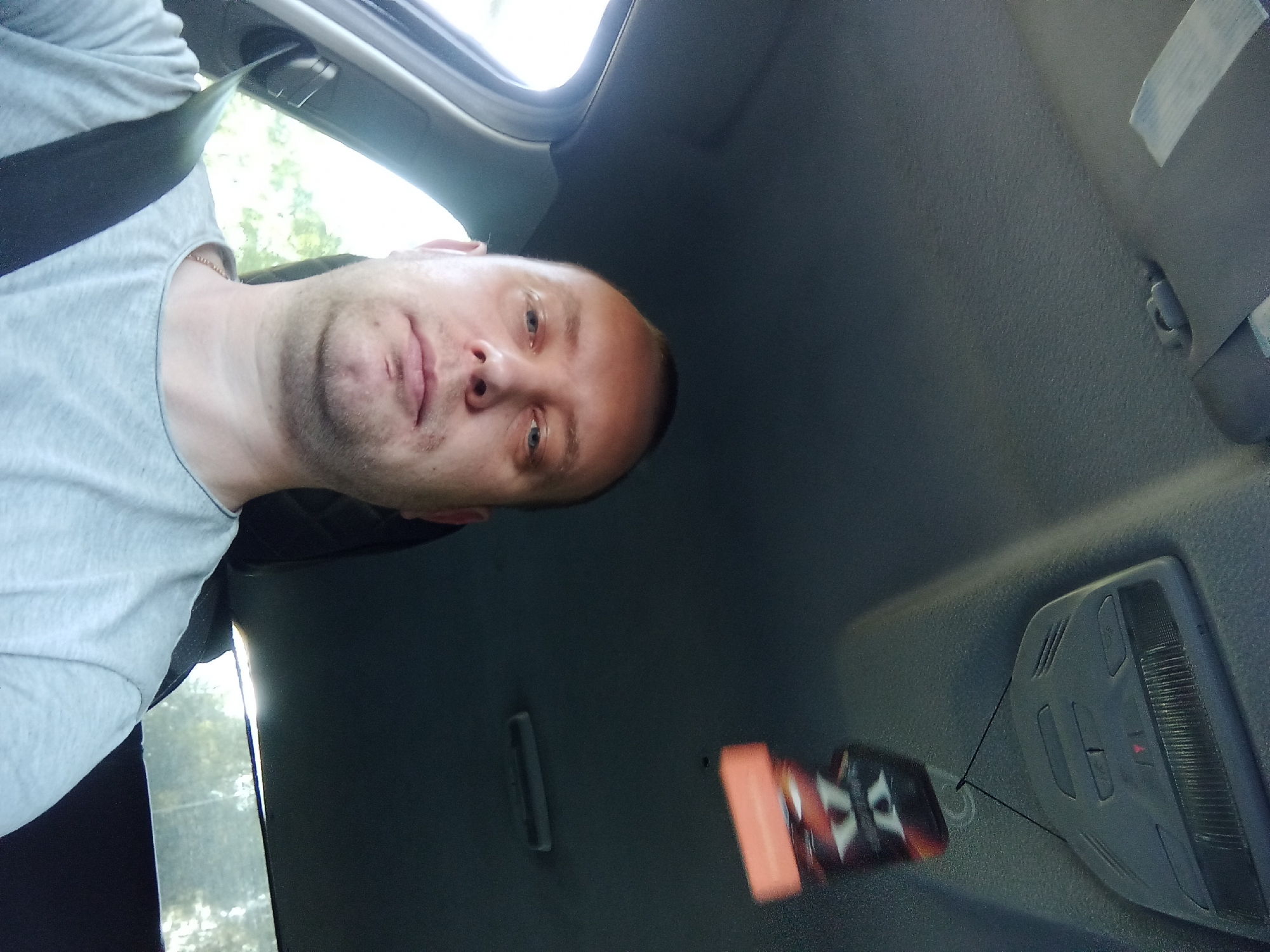 A bald white man wearing a grey t-shirt and a black seatbelt is sitting in the driver's seat of a car. He is looking directly at the camera. The interior of the car, including the dashboard, roof, and a car air freshener, is visible. The image was taken from inside the car, looking forward. The background outside the car appears to be a natural environment with trees. The location is Буйніцкі сельскі Савет, Belarus.