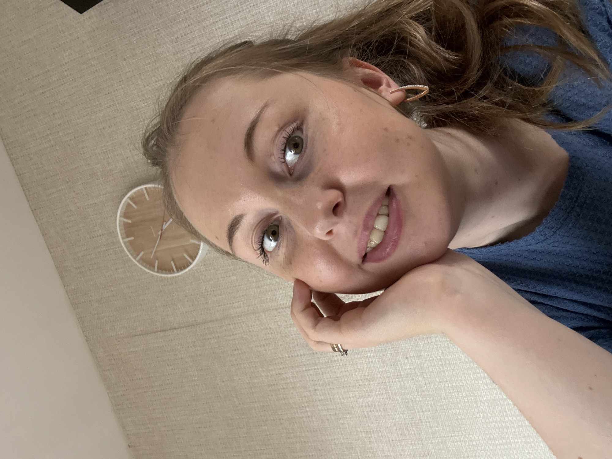The image is a close-up, rotated selfie of a young woman with light brown hair. She is looking at the camera with a slight smile. Her left hand is resting on her cheek. She is wearing a blue top and a small ring on her right hand. She has noticeable freckles on her face and arms. In the background, a light-colored textured surface is visible, possibly a wall or furniture. A round clock with a minimalist design is also partially visible on the textured surface to the left of the woman's face. The lighting suggests it could be daytime. There is no visible text in the image.