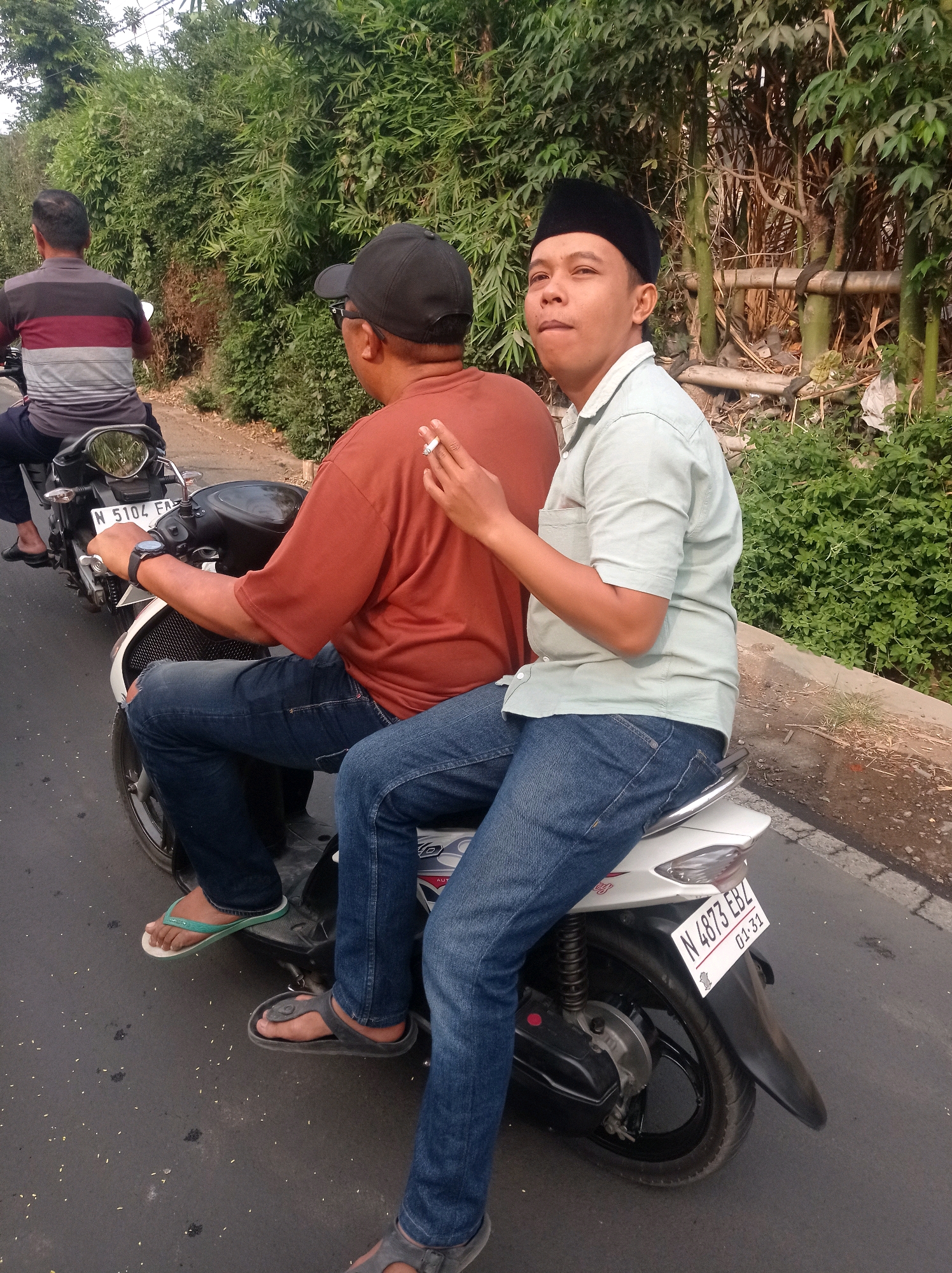 Two men are riding on a motorbike on a paved road in Pandanmulyo, Indonesia. The man in front is wearing a light blue short-sleeved collared shirt and blue jeans. He is looking at the camera and has a cigarette in his right hand, which is resting on the shoulder of the man behind him. The man behind him is wearing a rust-colored t-shirt and blue jeans. He is wearing a black skullcap and a black baseball cap. The motorbike is white and has a license plate that reads "N 4873 EBZ". There is lush green vegetation on the right side of the road, and a bamboo fence. Another motorbike with a man in a striped shirt is visible in the background on the left. The weather appears to be sunny.