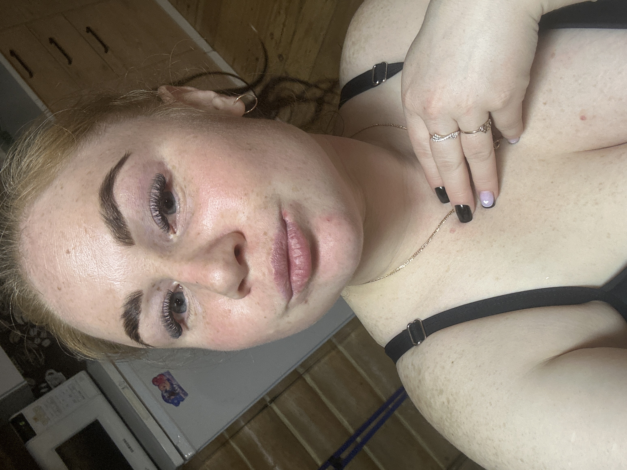 A close-up, rotated selfie shows a fair-skinned person with freckles, appearing to lie down with their head tilted. They have reddish-blonde hair, dark eyebrows, and eyelash extensions. They are wearing what appears to be a black garment with straps. The person's right hand is visible with multiple rings and nails painted with black and pale purple polish. A thin gold necklace is around their neck. The background shows a white refrigerator and a microwave. A wooden floor and cabinet are visible in the background. It is impossible to confirm the location is the city of Chaikovsky, Russia from the visual information provided.