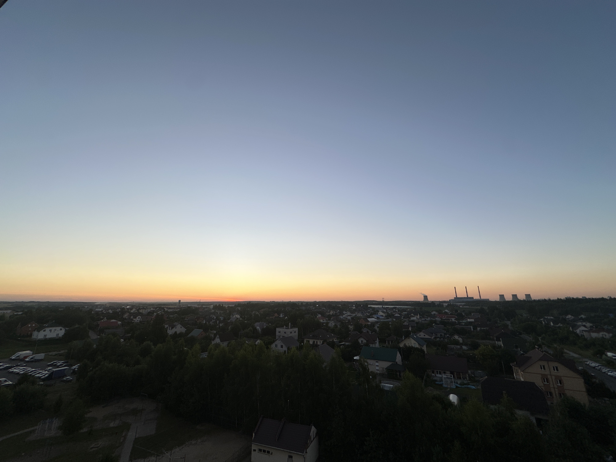 The image captures a panoramic view of a suburban area at dusk, likely on the outskirts of Шчомысліцкі сельскі Савет, Belarus. The sky transitions from a pale blue at the top to a soft orange and yellow near the horizon, indicating the setting sun. In the foreground and midground, dense green trees obscure a portion of the view. Dotted amongst the trees are numerous houses with pitched roofs, characteristic of a residential area. A parking lot filled with vehicles is visible on the left side of the image. In the distance, a cluster of industrial structures, including several tall cooling towers emitting faint plumes of smoke, stands against the twilight sky, suggesting the presence of a power plant or industrial complex. The overall scene depicts a tranquil, rural-industrial landscape at the end of the day.