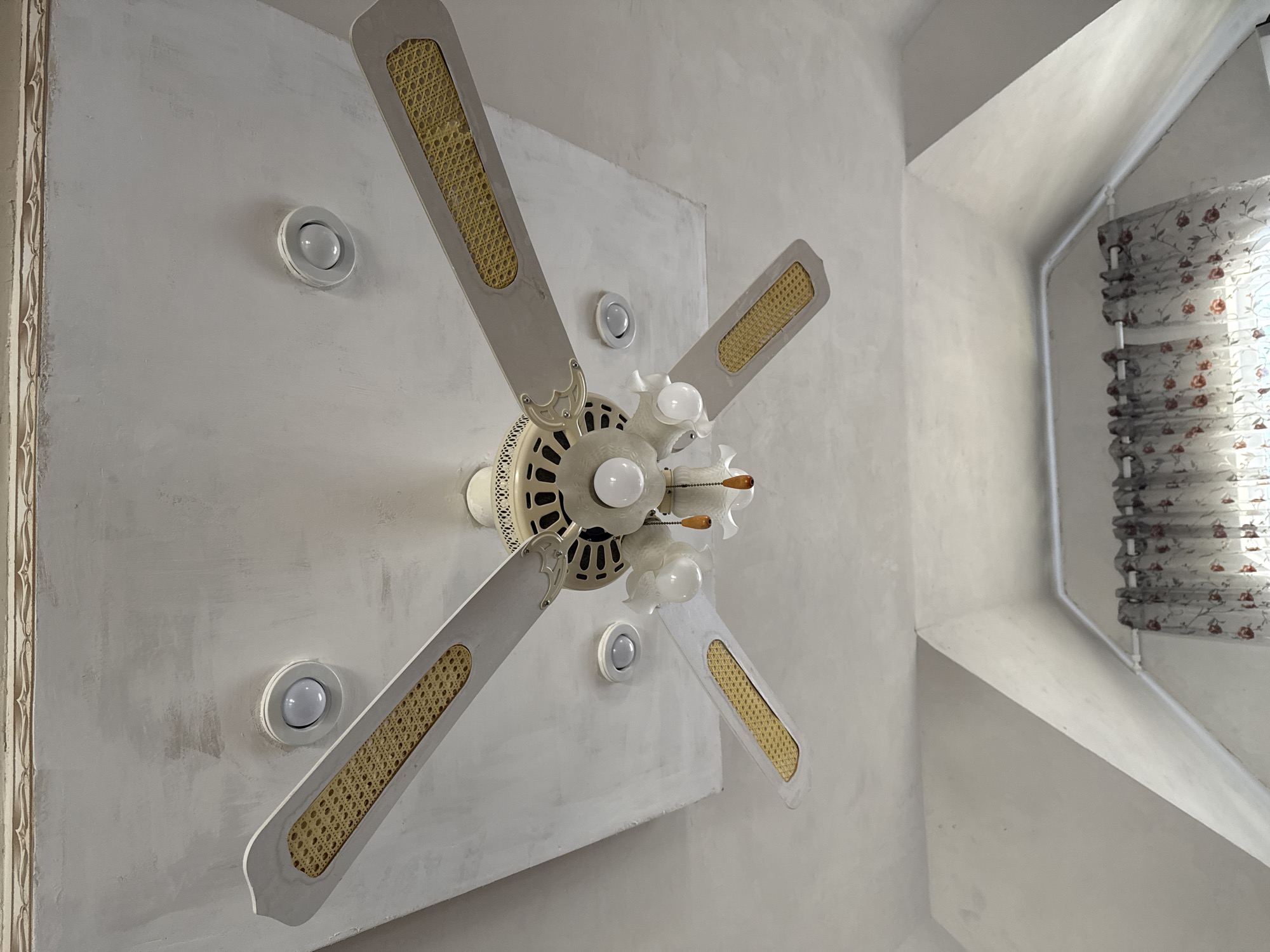 The image shows a ceiling fan with four blades, each having a wicker-like pattern, centered within a rectangular recess in the ceiling. The fan has a central light fixture with multiple bulbs. Recessed lighting fixtures are spaced around the fan within the rectangular recess. A window with a patterned shade is visible in the upper right corner of the image, suggesting a room interior. The style of the architecture and interior design appears dated. The location is unknown and there is no information to suggest this photo was taken in the city of Адам, Russia.