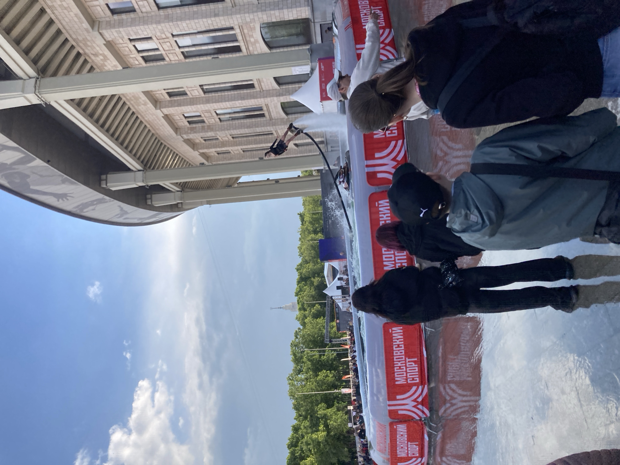 A public water-jet stunt performance event is taking place in the city of Moscow, Russia. A building with rows of windows can be seen in the background. A water-jet performer is performing a stunt in the air above the water, propelled by the water jets. A crowd of people is watching the performance from behind a red and white safety barrier with "МОСКОВСКИЙ СПОРТ" written on it. The sky is blue with some white clouds. The spire of one of the Seven Sisters skyscrapers is visible in the background.