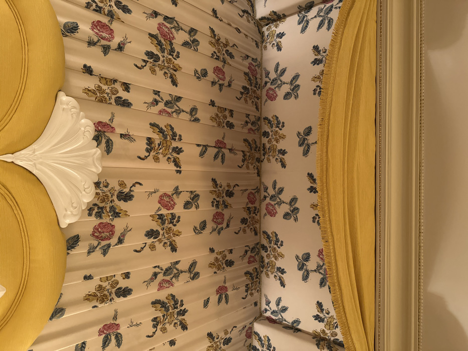 The image captures the corner of a room with floral wallpaper, predominantly roses and foliage in shades of red, green, and cream. A bright yellow, tufted armchair with a white ornamental headrest occupies the left side. A thick, yellow curtain with fringed edges hangs along the corner where the wallpapered wall meets a white-framed wall. The floral pattern extends to the ceiling, creating an enclosed, cozy feel. The composition is a close-up shot, showcasing the textures and patterns of the room's interior design.