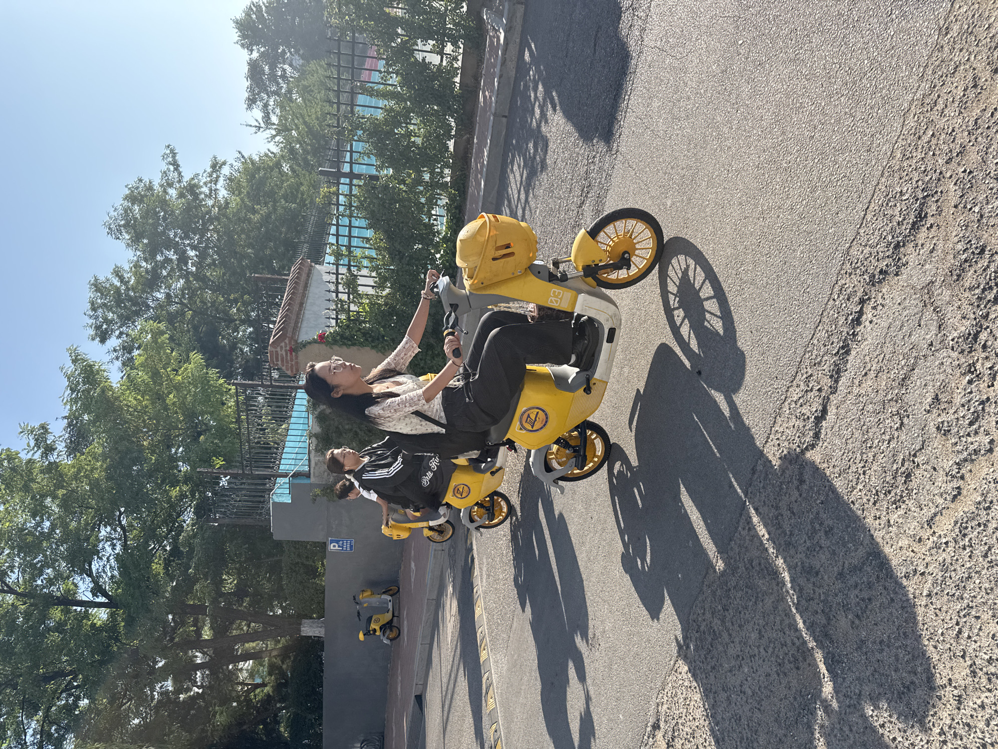 Two women are riding yellow electric scooters on a paved road in Zhongtun, China. The woman in the foreground, wearing glasses, a white lace top, and black pants, is looking forward. The woman behind her, wearing a black jacket with white stripes on the sleeves and black pants, is also looking forward. Both scooters have baskets in the front and a number plate that reads "03". The scene is bathed in sunlight, casting long shadows on the road. In the background, there are trees, a fence, and a building with a blue structure. A parked yellow scooter is visible on the sidewalk in the distance, next to a blue and white parking sign.