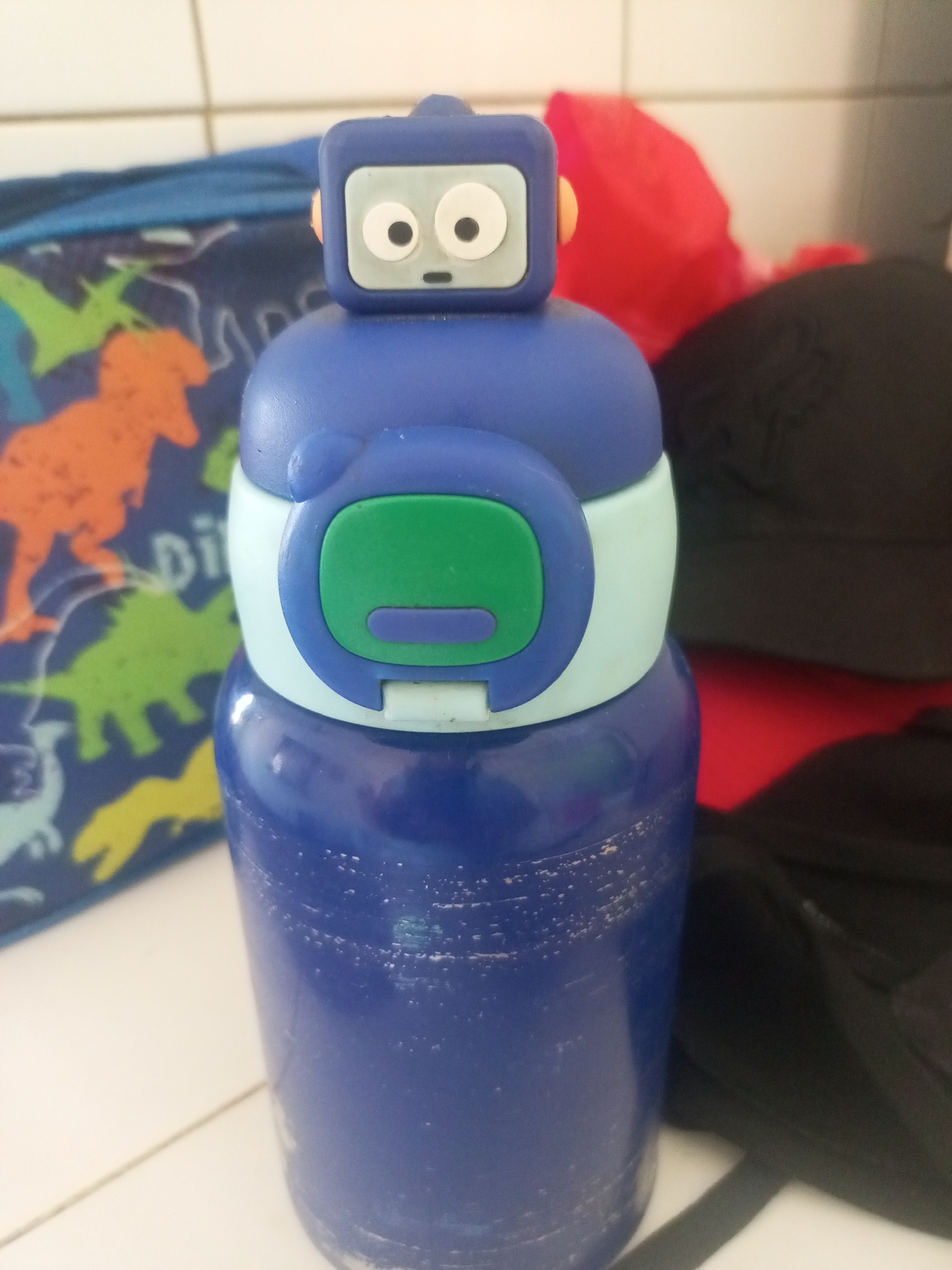 A blue robot-shaped water bottle is the central focus of this image. It stands upright on a white surface, possibly a tiled countertop. The robot's head is blue and square with white circular eyes and a small grey mouth. The body of the robot is composed of a light blue section and a larger blue section that forms the main body of the bottle. A green rectangular button with a purple rectangular detail is present on the light blue part of the robot's body.
In the background, to the left of the bottle, a blue bag with dinosaur prints in orange, green, and yellow is partially visible. Behind the robot, and slightly to the right, a bright red object, possibly fabric or plastic, is visible, along with a dark-colored object, likely a bag or backpack. The overall environment appears to be indoors, with a white tiled wall in the background. The lighting suggests it is daytime. There is no indication of the location being the city of La Tagua, Venezuela.