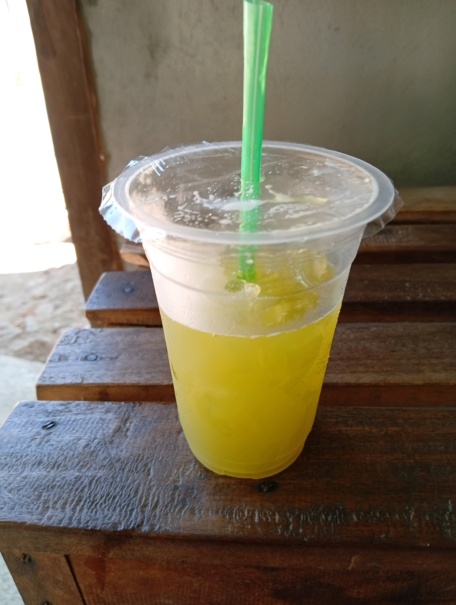 A close-up, eye-level shot shows a clear plastic cup filled with a bright yellow liquid, likely juice, with ice cubes visible. A vibrant green plastic straw protrudes from the top of the cup, which is sealed with a clear plastic lid. The cup rests on a wooden surface, possibly a table or bench, composed of horizontal planks. The background is blurred, but suggests an outdoor setting with a wall and sunlight casting shadows, possibly indicating the location is in Sukowetan, Indonesia. The overall lighting suggests a bright, daytime scene.