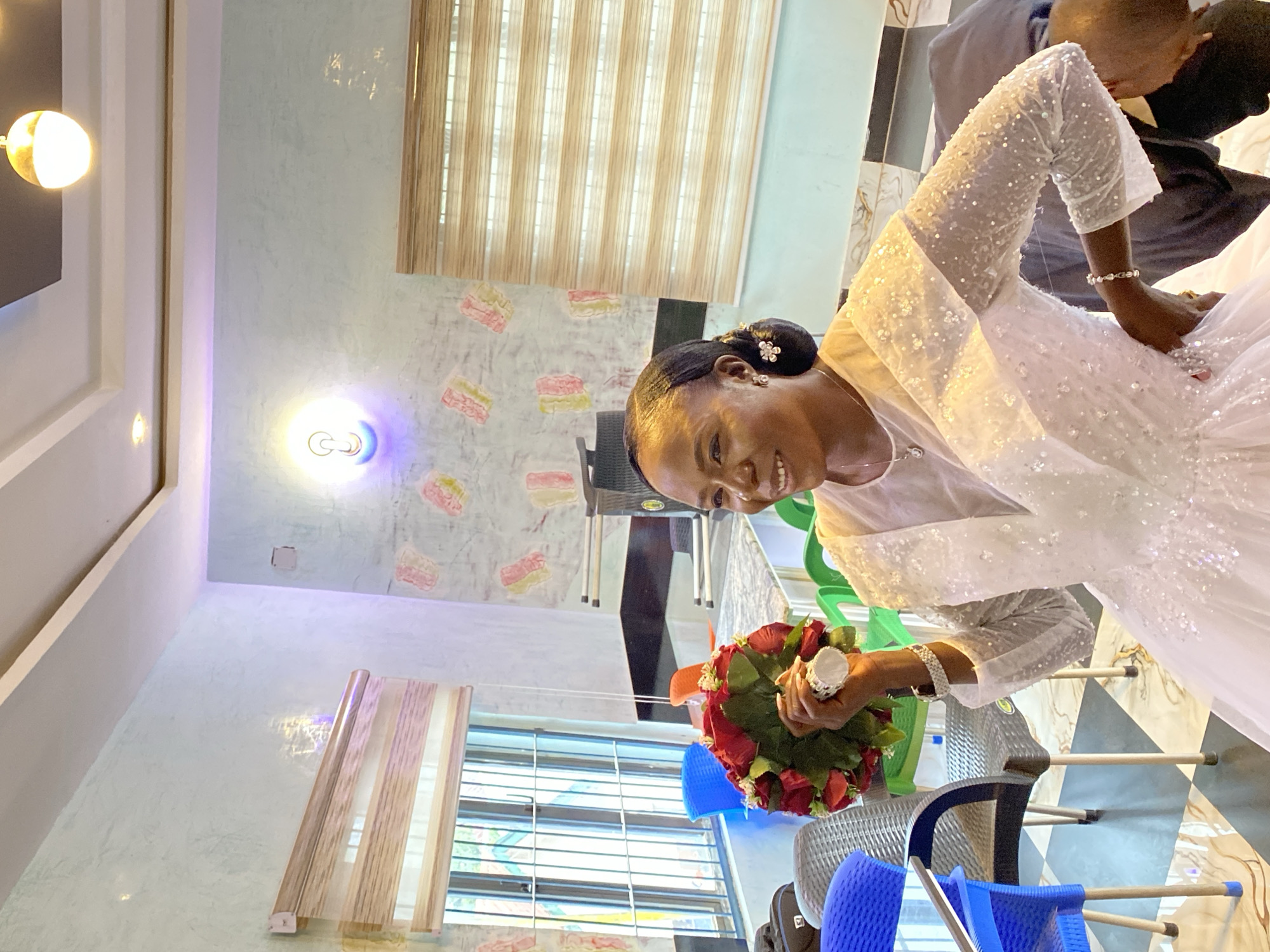 A bride stands in a room in Kuru Dungus, Nigeria, holding a bouquet of red roses. She wears a white wedding dress with beaded long sleeves. The room has blinds on the windows, patterned wallpaper, and overhead lighting. Chairs are visible in the background. A man in dark clothing stands behind the bride, interacting with her.