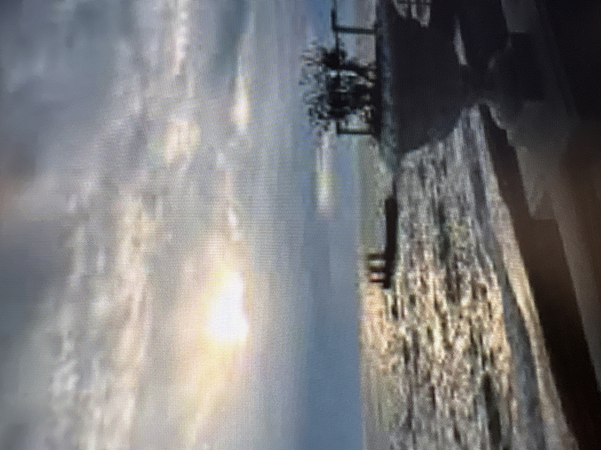The image depicts a hazy, sunlit scene over a body of water, possibly a large river or lake, with a barge or boat in the distance. In the foreground, there's a blurred structure that appears to be a planter or decorative element with some foliage. The sky is cloudy, with the sun breaking through, creating reflections on the water. The overall impression is one of an outdoor setting, likely a waterfront area, with soft lighting and a somewhat muted color palette due to the atmospheric conditions. The image's low resolution and blurriness make it difficult to discern specific details about the location or any individuals. There is no indication that this scene is located in Moscow, Russia.