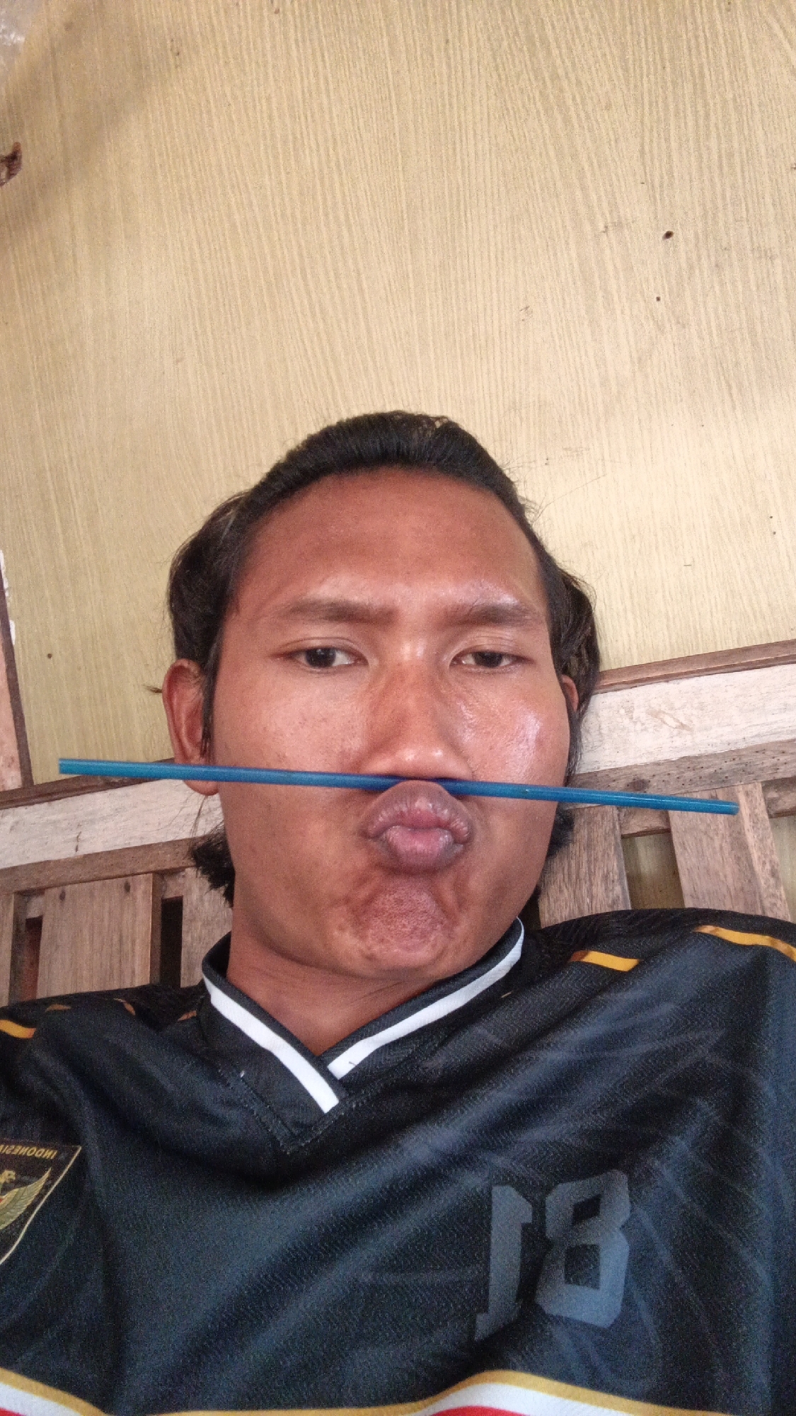 A medium shot captures a person with dark hair, a light brown complexion, and a short beard, holding a blue plastic straw horizontally across their mouth. The person is wearing a black jersey with white and yellow accents. They are seated on a wooden bench, with a textured yellow wall in the background. The lighting is natural and appears to be from an overhead source. The setting is indoors, possibly a home or a casual establishment, with the presence of wooden furniture suggesting a rustic or traditional environment.