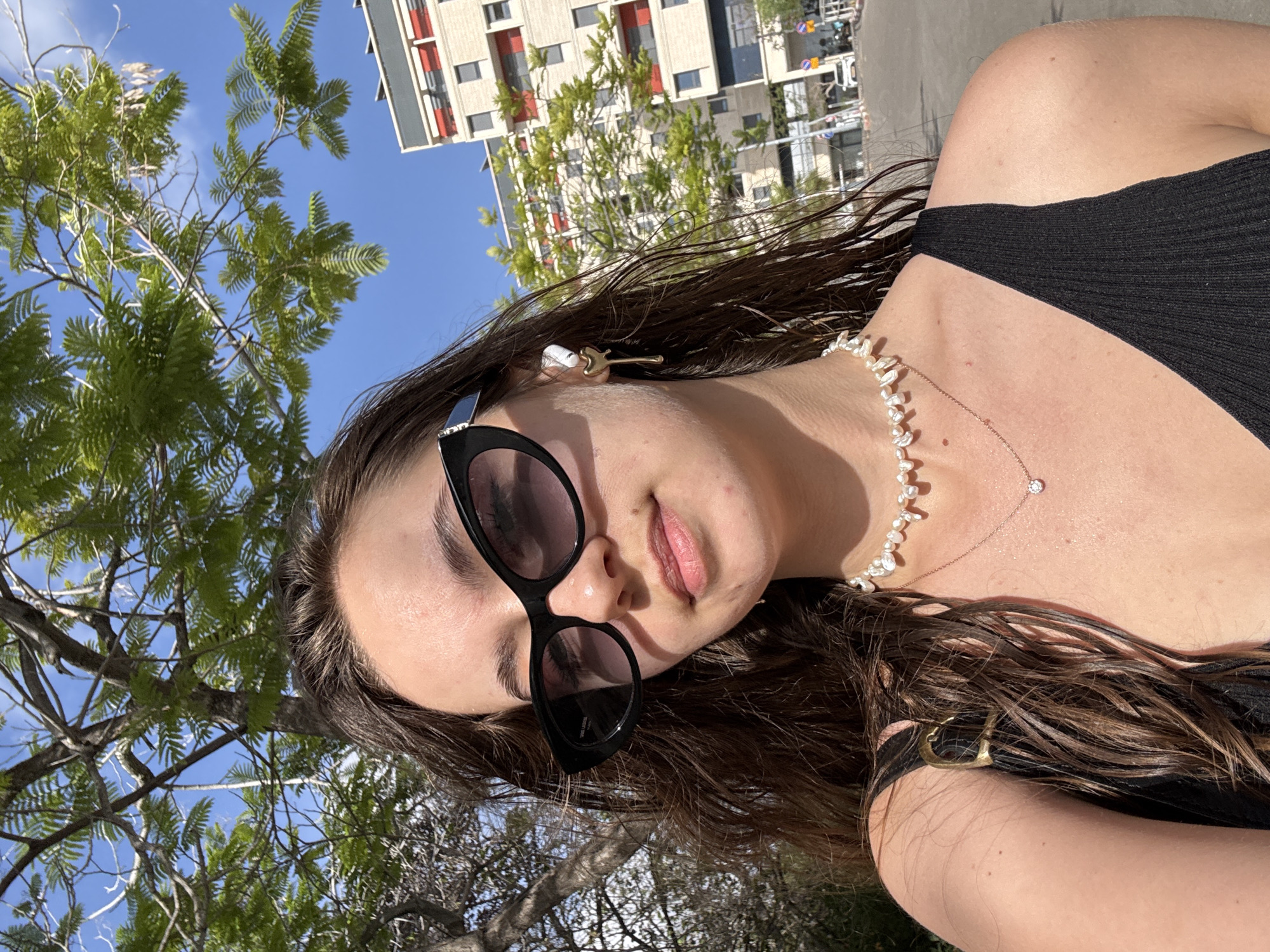 A young woman is pictured in an outdoor setting. She has fair skin, brown hair, and is wearing sunglasses, a pearl necklace, a black top, and a gold guitar-shaped hair clip. The background includes trees, a blue sky, and a building with a red accent. It's Tel-Aviv, Israel.