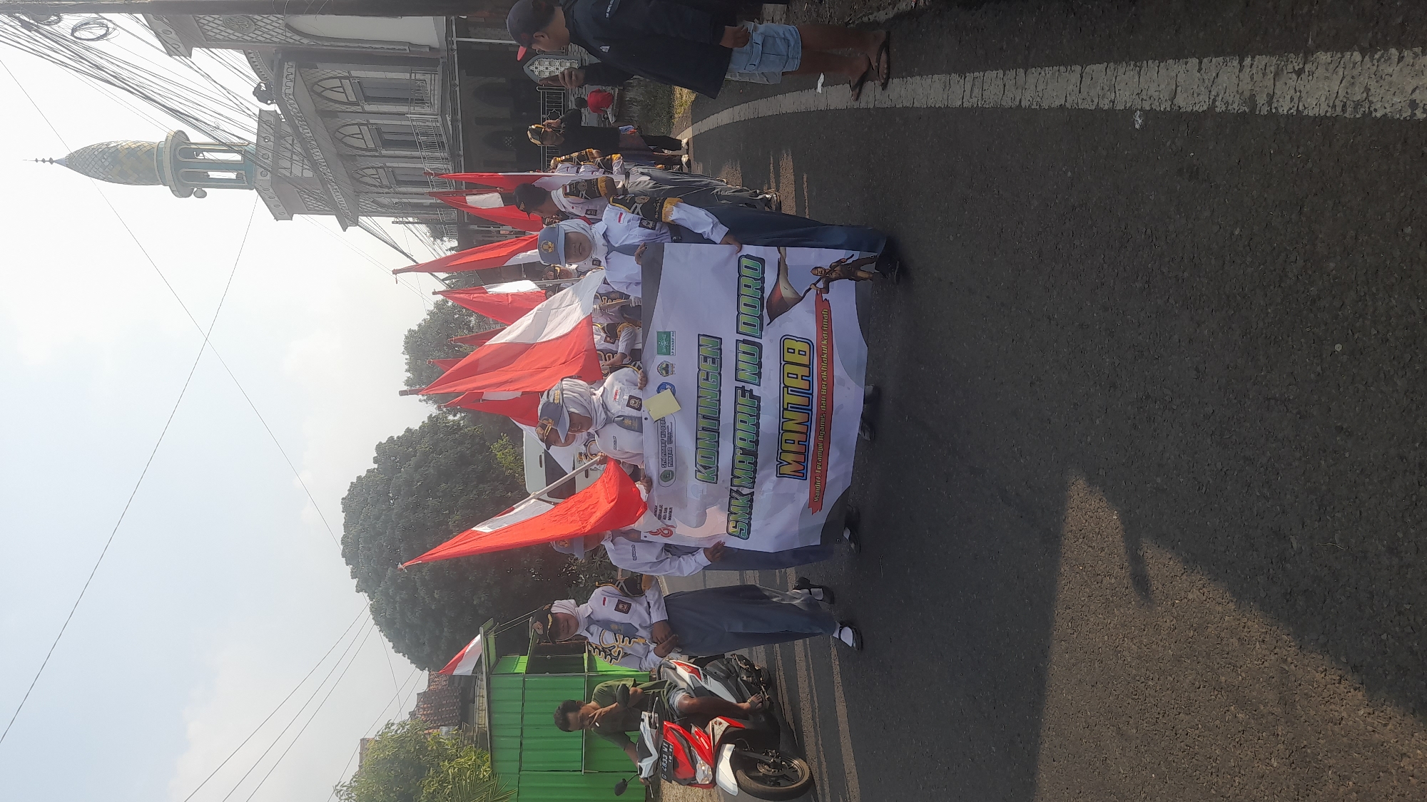 The image depicts a street scene in Lemahabang, Indonesia, during what appears to be a procession or parade. Several individuals, predominantly young women dressed in school uniforms consisting of white shirts, grey skirts, and headscarves, are walking in a line. They are holding numerous Indonesian flags, which are red and white. One group is carrying a large banner that reads "KONTINGEN SMK MA'ARIF NU DORO MANTAB" and below that, "Mandiri, Terampil, Ngamul, dan Berakhlakul Karimah." The banner also features an illustration of a person holding a flag.
In the background, a mosque with a distinctive dome and minaret is visible, suggesting the location is likely near a religious center. A man on a red scooter is also present, and other bystanders are observing the procession. The environment appears to be a paved road with trees and buildings lining the sides. The lighting indicates it is daytime, possibly in the morning or late afternoon. The presence of the flags and the school uniforms strongly suggests a patriotic or celebratory event related to the school.