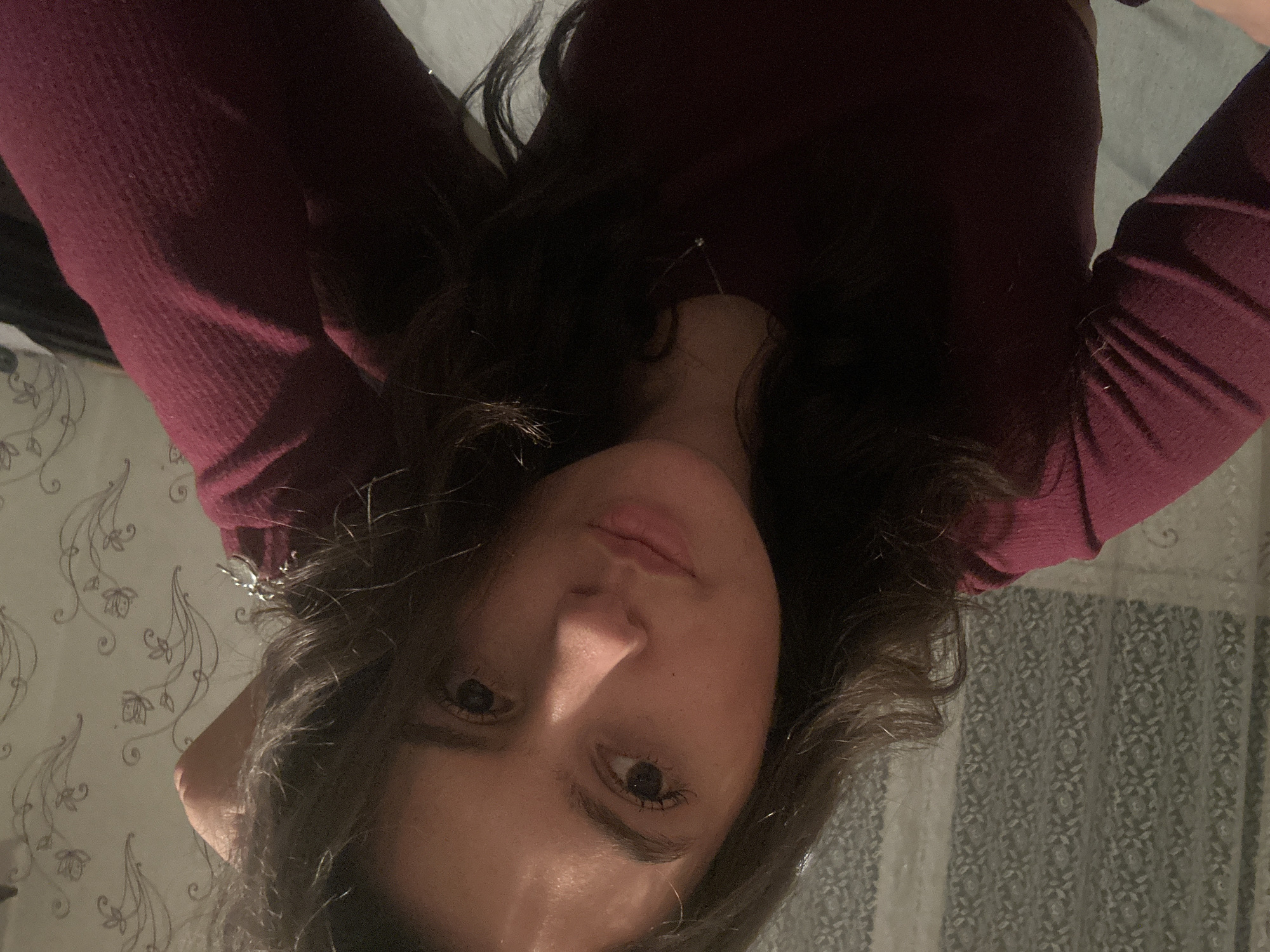 A woman with wavy, dark hair is seen in a low-angle, close-up selfie. She is wearing a long-sleeved, maroon-colored garment. Her face is the primary focus, with visible eyes, nose, and lips. The background is a patterned surface that may be wallpaper or fabric. The location is unspecified, Unknown, Unknown.