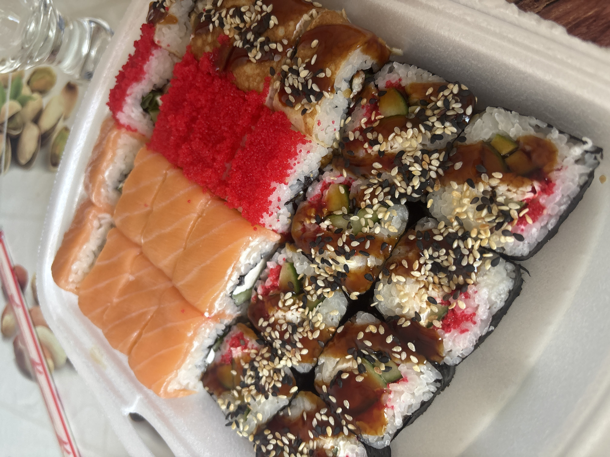 The image shows a takeout container of sushi. The sushi varieties include salmon rolls, red tobiko rolls, and rolls with eel, sesame seeds, cucumber, and sauce. The sushi is in a white styrofoam container. A straw and what appears to be pistachios are visible on the side. The location is not specified, but the prompt indicates it is in Chaikovsky, Russia.