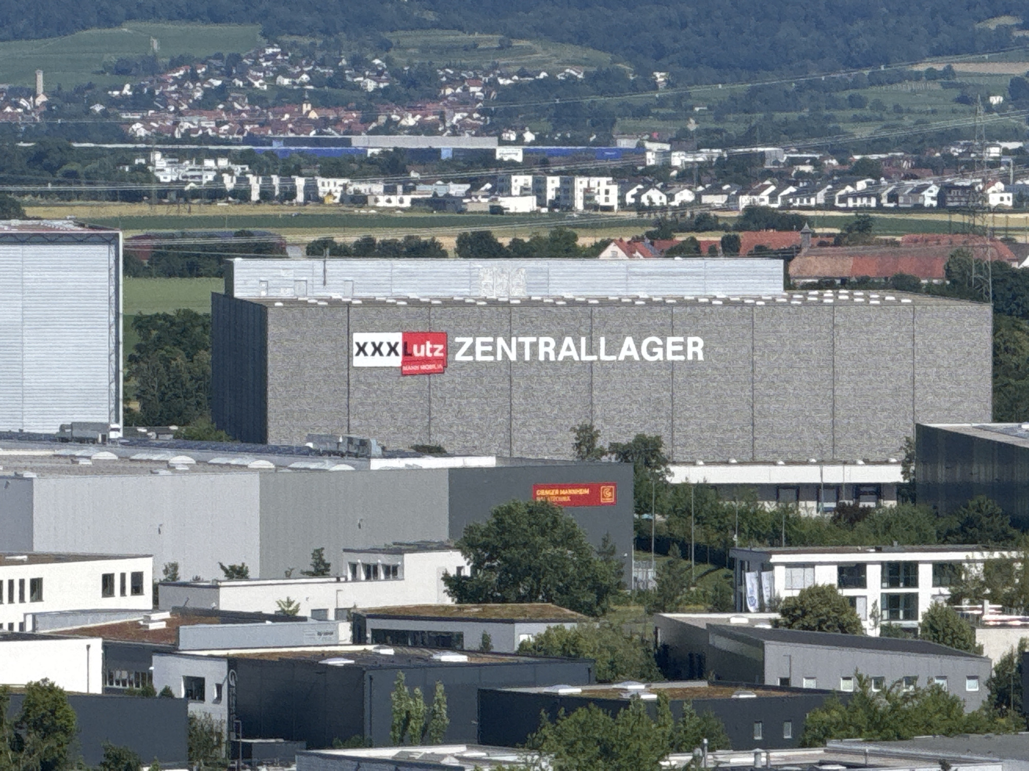 The image is an aerial, telephoto view of an industrial or commercial area, likely on the outskirts of Mannheim, Germany. The most prominent structure is a large, rectangular building with the text "XXX LUTZ ZENTRALLAGER" emblazoned on its side. The text is in white, with "XXX LUTZ" on a red background. The building appears to be a distribution or storage facility.
Surrounding this main building are other industrial buildings of varying sizes and designs, many with flat roofs. Some of these buildings are grey, others are white, and some have green roofs. One building in the mid-ground has a red rectangular sign with white text that appears to say "DRAGER HAUPTEN" and a logo.
In the background, a more densely populated area with residential buildings, possibly a suburb or part of the city, is visible. These buildings are a mix of white and red roofs. Further in the distance, rolling green hills form the backdrop to the scene. The sky is clear and the lighting suggests it is daytime, likely late morning or early afternoon. There are no people or vehicles clearly visible in the image. The overall impression is of a functional industrial zone.