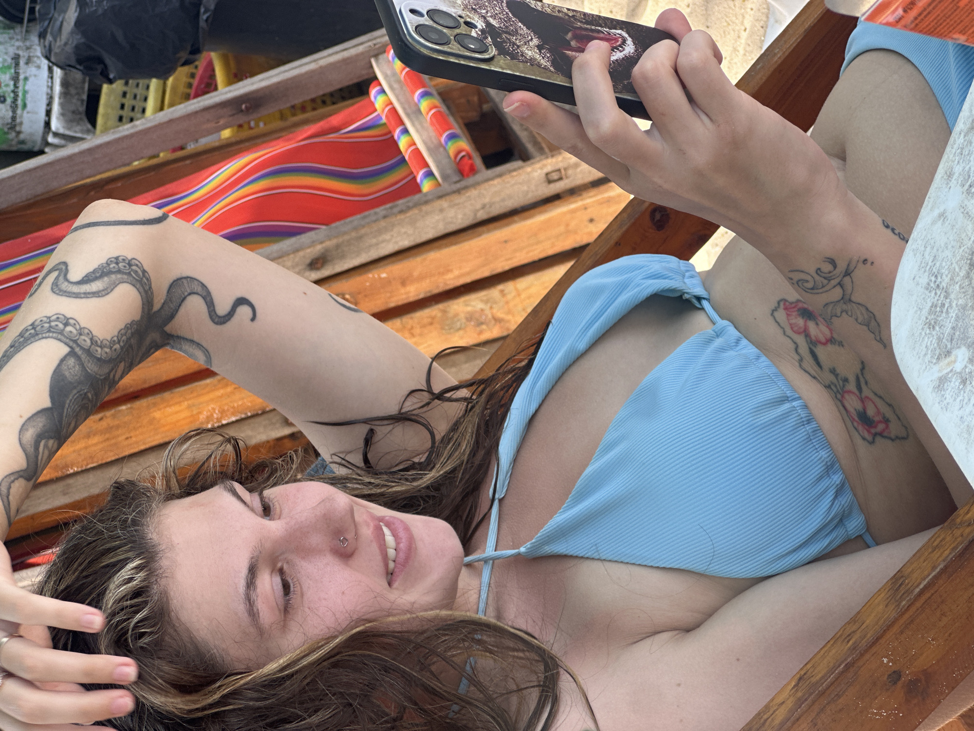 A woman lies on a wooden lounger, apparently in Baan Chaweng, Thailand. She is wearing a light blue bikini top and holding a smartphone with an alligator or crocodile design case. The woman has an octopus tattoo on her left arm and a floral tattoo on her right arm. She is smiling and looking at the phone. The lounger has colorful striped cushions behind her.