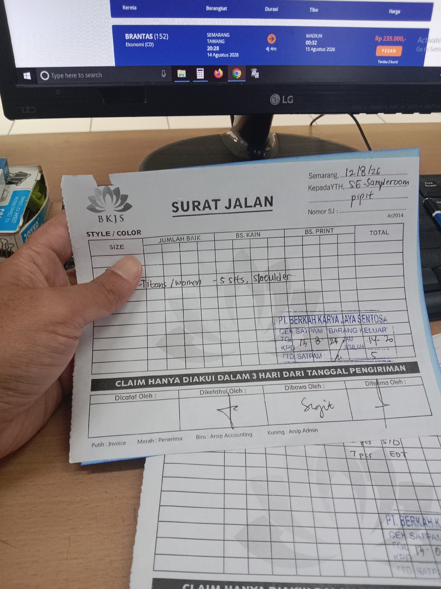 The image shows a close-up view of a person's hands holding a document that appears to be a "SURAT JALAN" (delivery order) or shipping document. The document has the logo "BKJS" at the top left. It contains various fields for information such as "STYLE/COLOR," "SIZE," "JUMLAH BAIK," "BS. KAIN," "BS. PRINT," and "TOTAL." Below this table, there's a section for "CLAIM HANYA DIAKUI DALAM 3 HARI DARI TANGGAL PENGIRIMAN" (Claims are only recognized within 3 days of the delivery date). Fields for "Dicatat Oleh" (Recorded By), "Diketahui Oleh" (Known By), "Dibawa Oleh" (Carried By), and "Diterima Oleh" (Received By) are visible.
In the upper part of the image, a computer screen is partially visible, displaying a website with information about train travel, including details for "BRANTAS (152) Ekonomi (CD)" from SEMARANG to MADIUN with a departure time of 00:32 on 15 Agustus 2026 and a price of Rp 235.000,-. A portion of a keyboard is also visible.
There is handwritten text on the "SURAT JALAN" document. Specifically, it reads "Semarang, 12/18/26" at the top, followed by "Kepada YTH, S.G. Sameroom" and then "pipit" written below it. Further down, there is a stamp that reads "PT. BERKAH KARYA JAYA SENTOSA" with additional text like "CEK SATPAM BARANG KELUAR." Another handwritten entry appears to be a date and some numbers.
The scene appears to be an indoor setting, likely an office or a desk area, given the presence of the computer, keyboard, and documents. The lighting suggests it is daytime. There are no people fully visible, only hands holding the document. The general context suggests administrative or logistical work related to deliveries or claims. The mention of "Semarang" on the document and on the computer screen indicates a connection to this city in Indonesia.